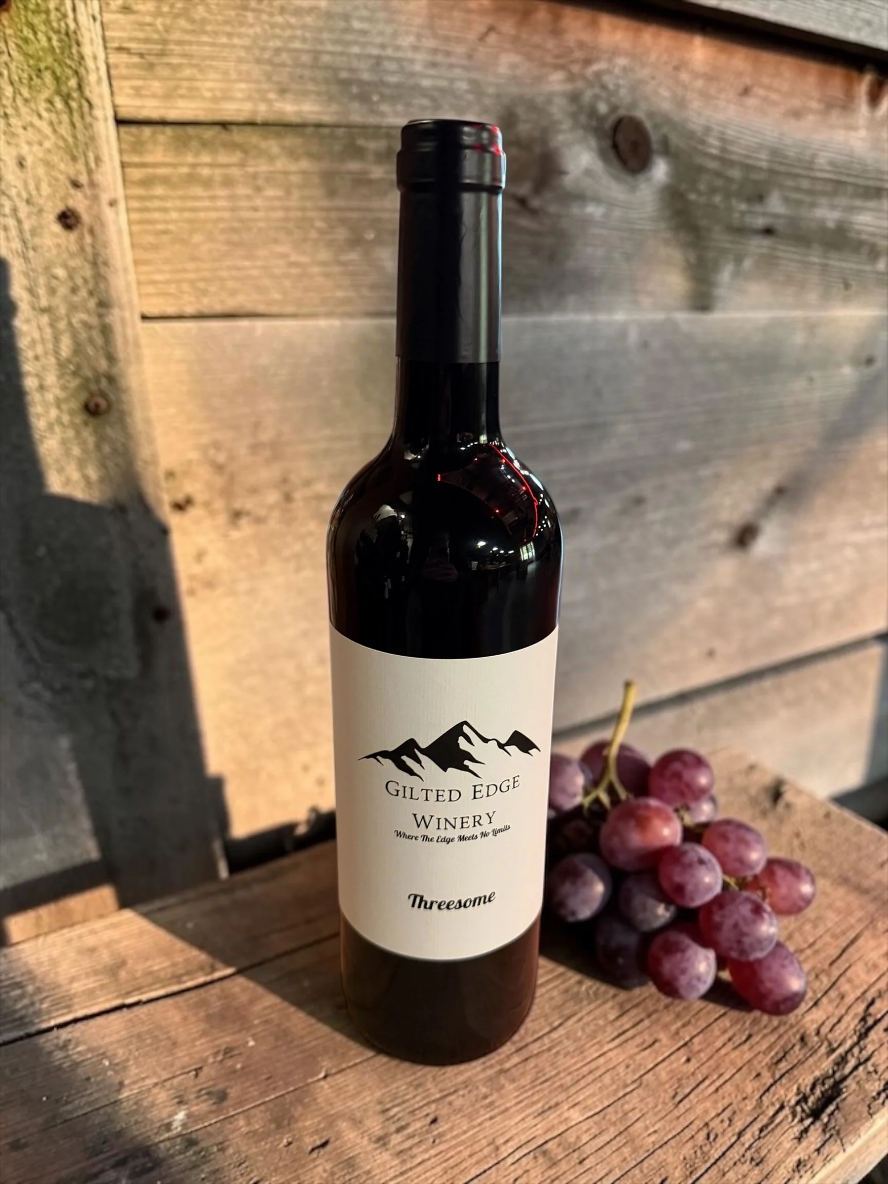 A bottle of wine from Gilted Edge Winery on a wooden surface with a bunch of red grapes next to it.