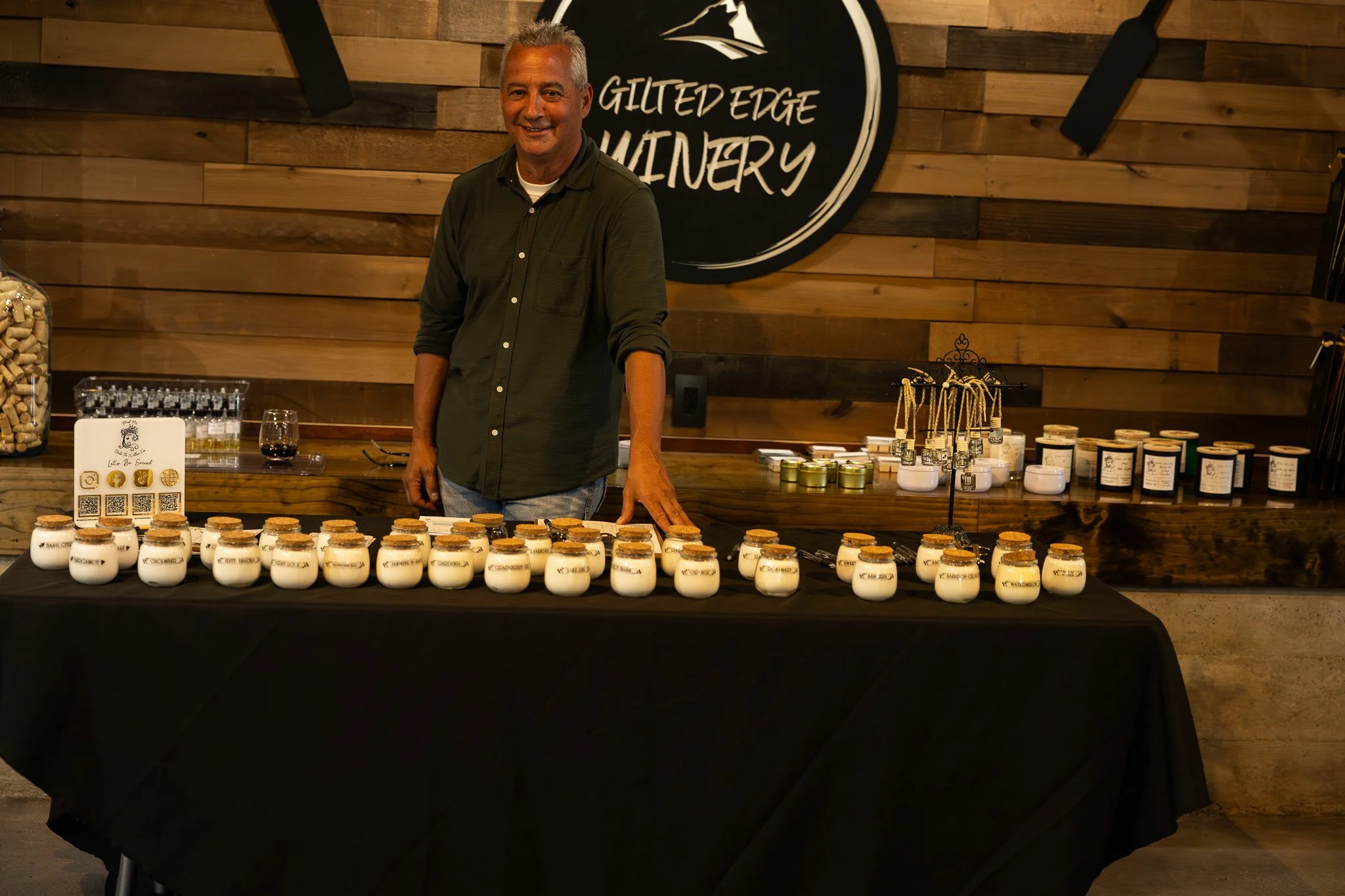 A man standing behind a table with small labeled jars, in a bakery or shop setting, with a wooden wall and a round sign that reads 'Gilted Edge Winery' behind him.