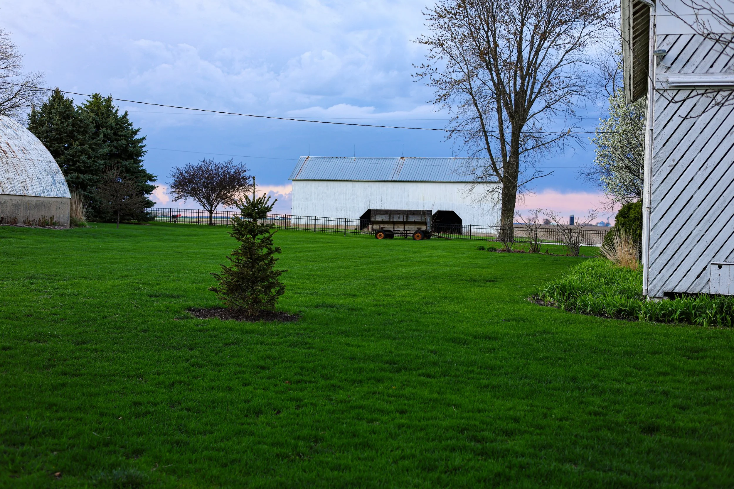 A rural scene with green grass, trees, a white barn with a metal roof, and a cloudy sky
