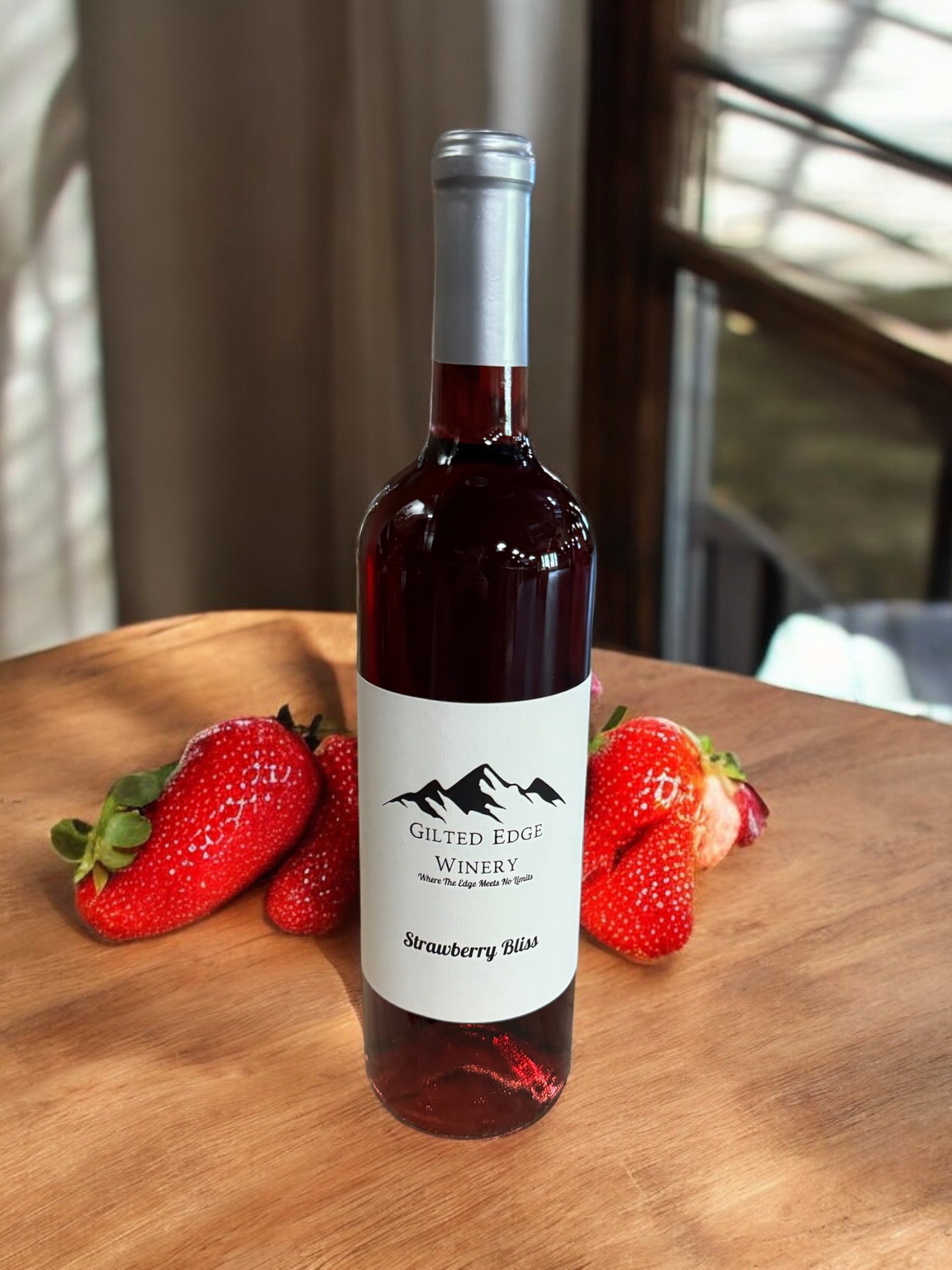 A bottle of Strawberry Bliss wine from Gilt Edge Winery, placed on a wooden surface with fresh strawberries around it. The wine appears to be a dark red color.