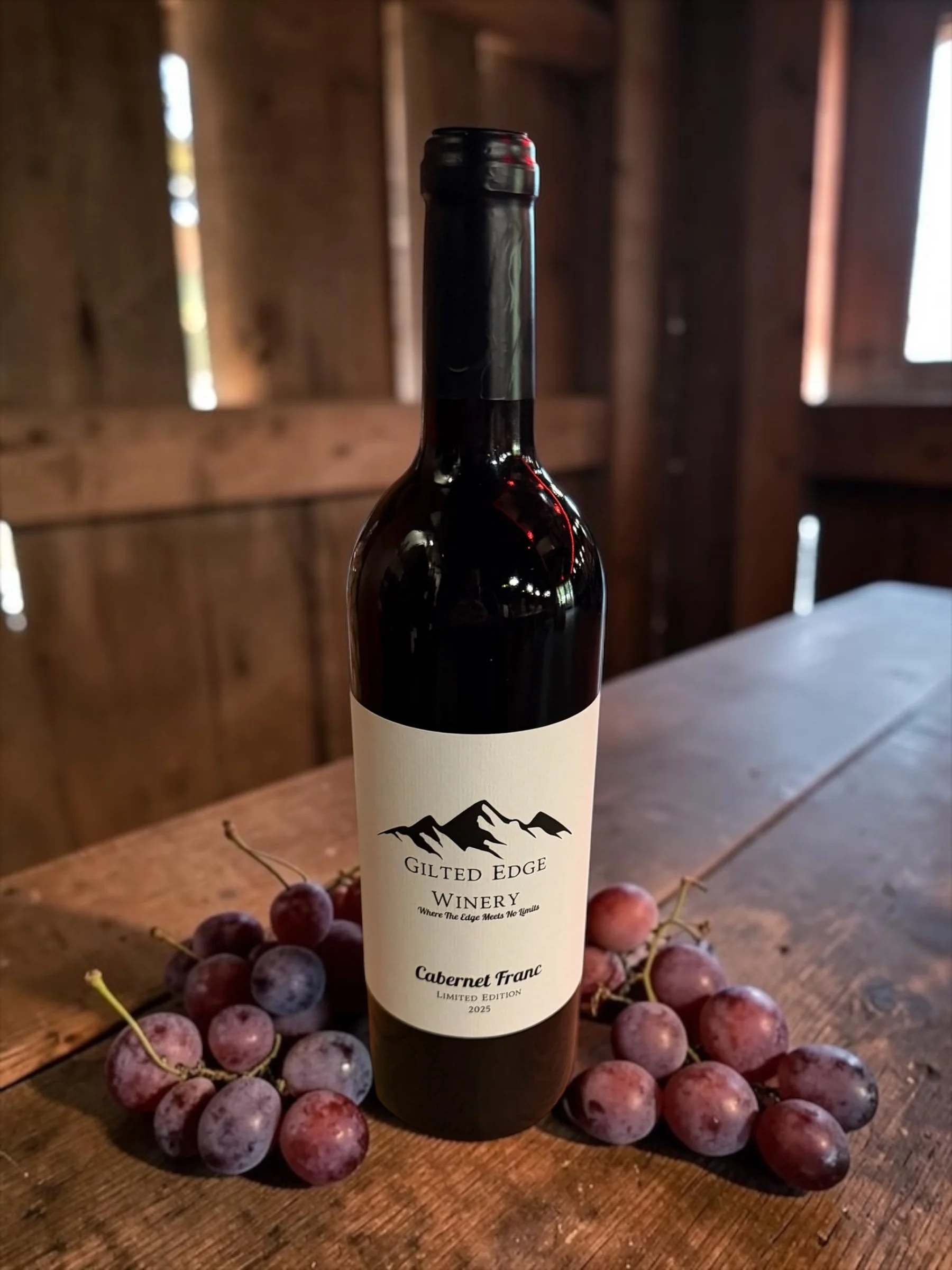 A bottle of Gilt Edge Winery Cabernet Franc wine placed on a wooden surface surrounded by red and purple grapes with a barn-like wooden interior in the background.