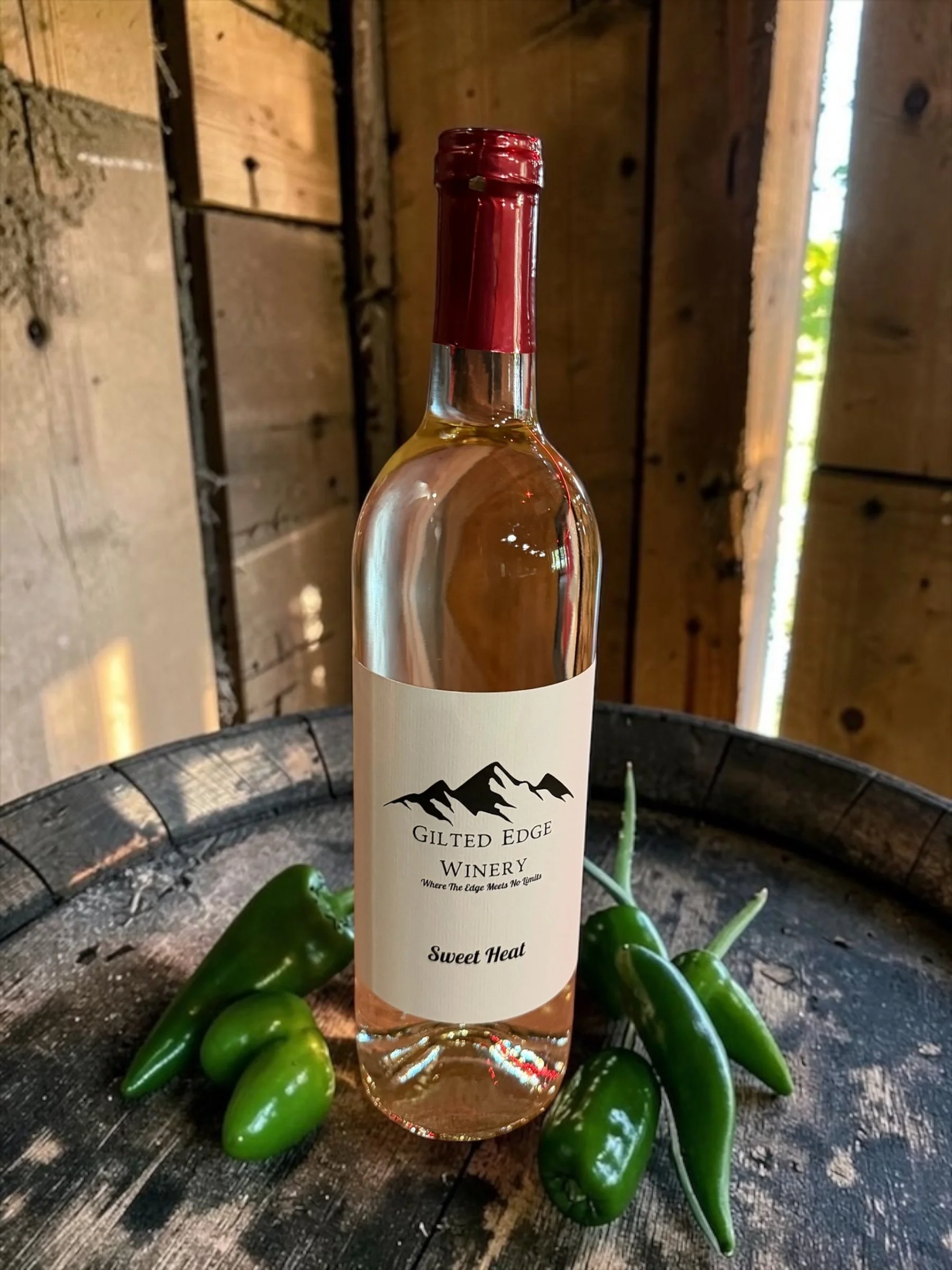 A bottle of wine from Gilded Edge Winery with a mountain logo and the label reading 'Sweet Heat,' surrounded by jalapeño peppers on a rustic wooden surface.