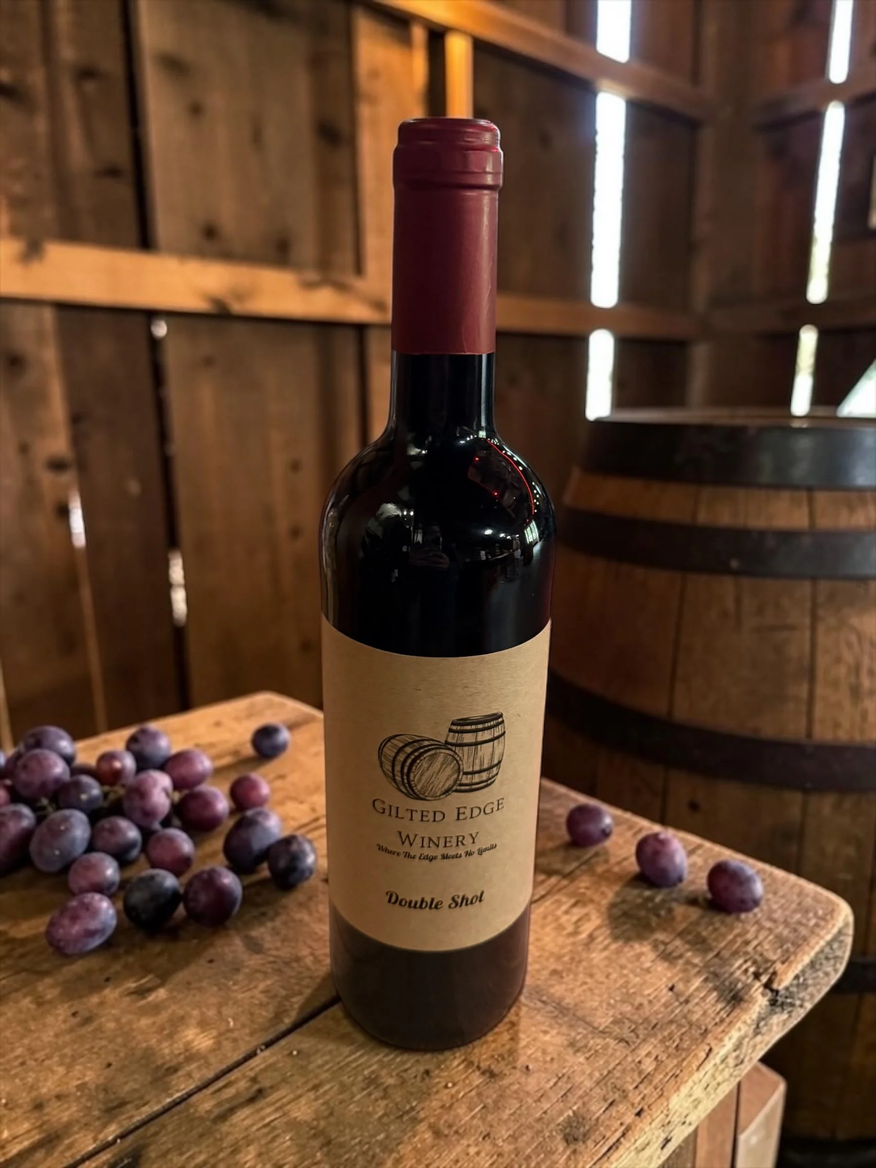 A bottle of red wine from Gilded Edge Winery with a label that says 'Double Shot,' placed on a wooden table with scattered grapes and the background of a rustic winery interior.