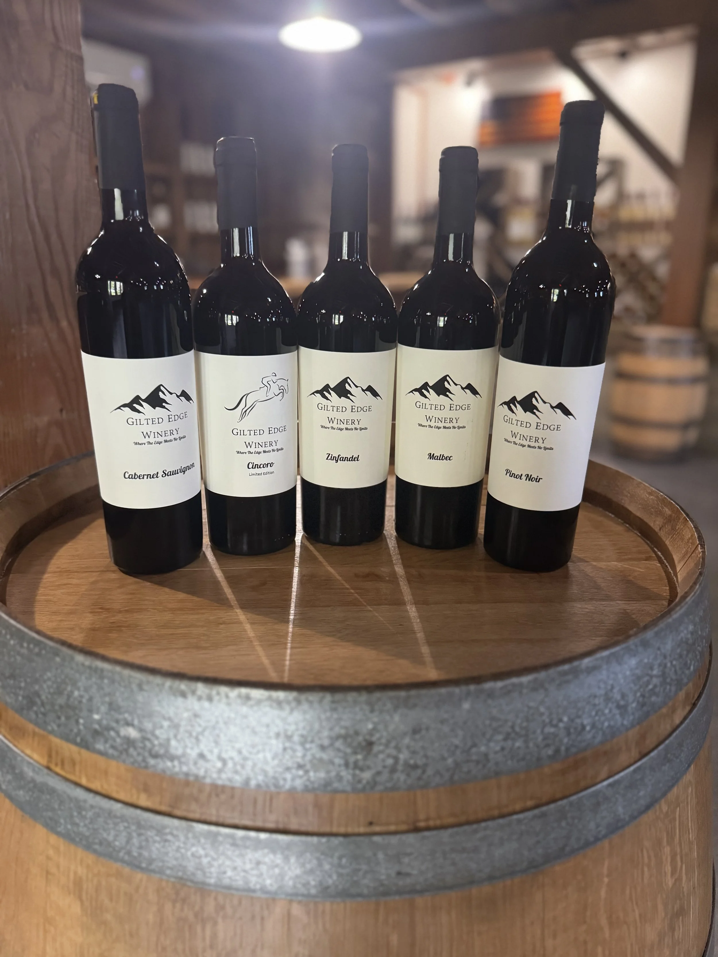 Five bottles of wine with labels from Gilted Edge Winery, including Cabernet Sauvignon, Cincoro limited edition, Zinfandel, Malbec, and Pinot Noir, displayed on a wooden surface inside a wine barrel.