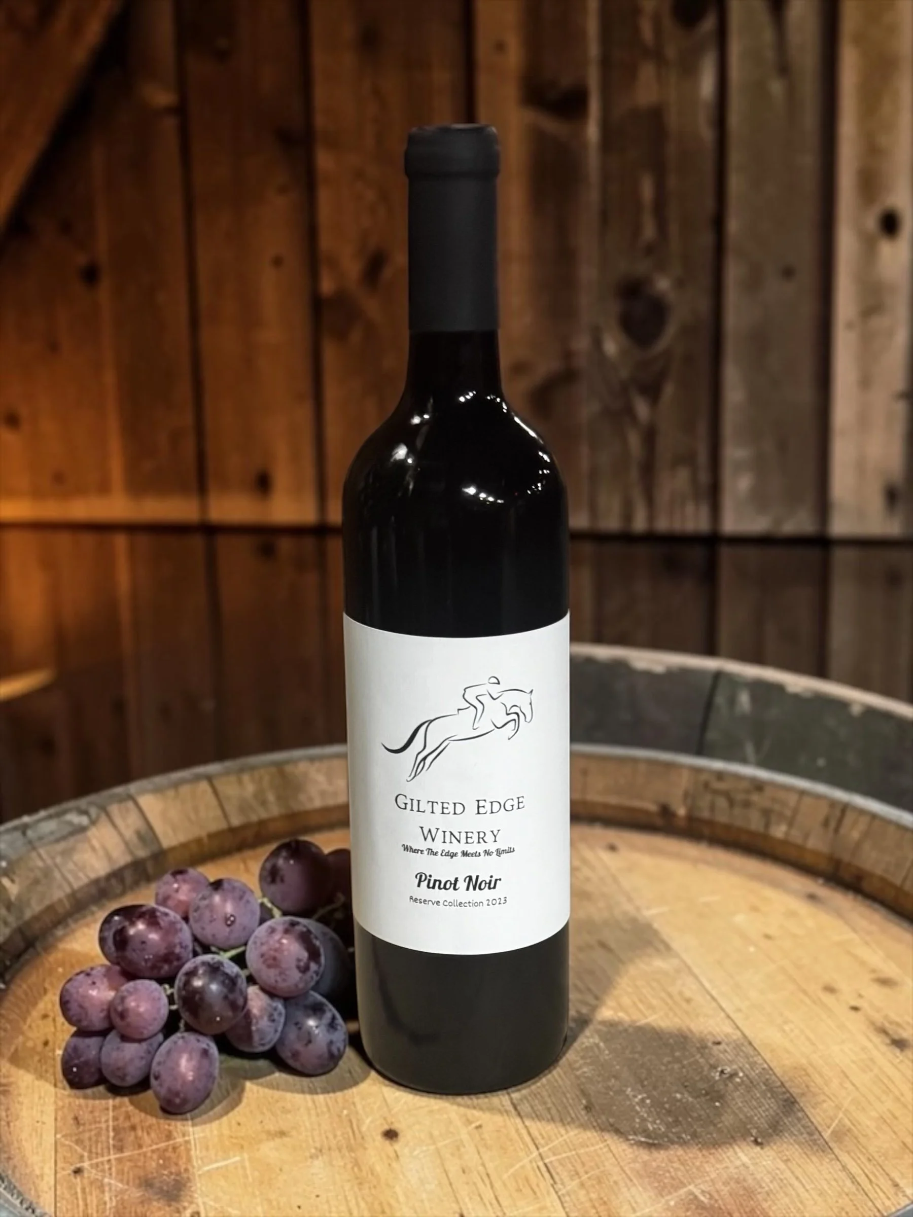 A bottle of Gilded Edge Winery Pinot Noir wine on a wooden table, with purple grapes in a basket nearby, against a wooden wall background.