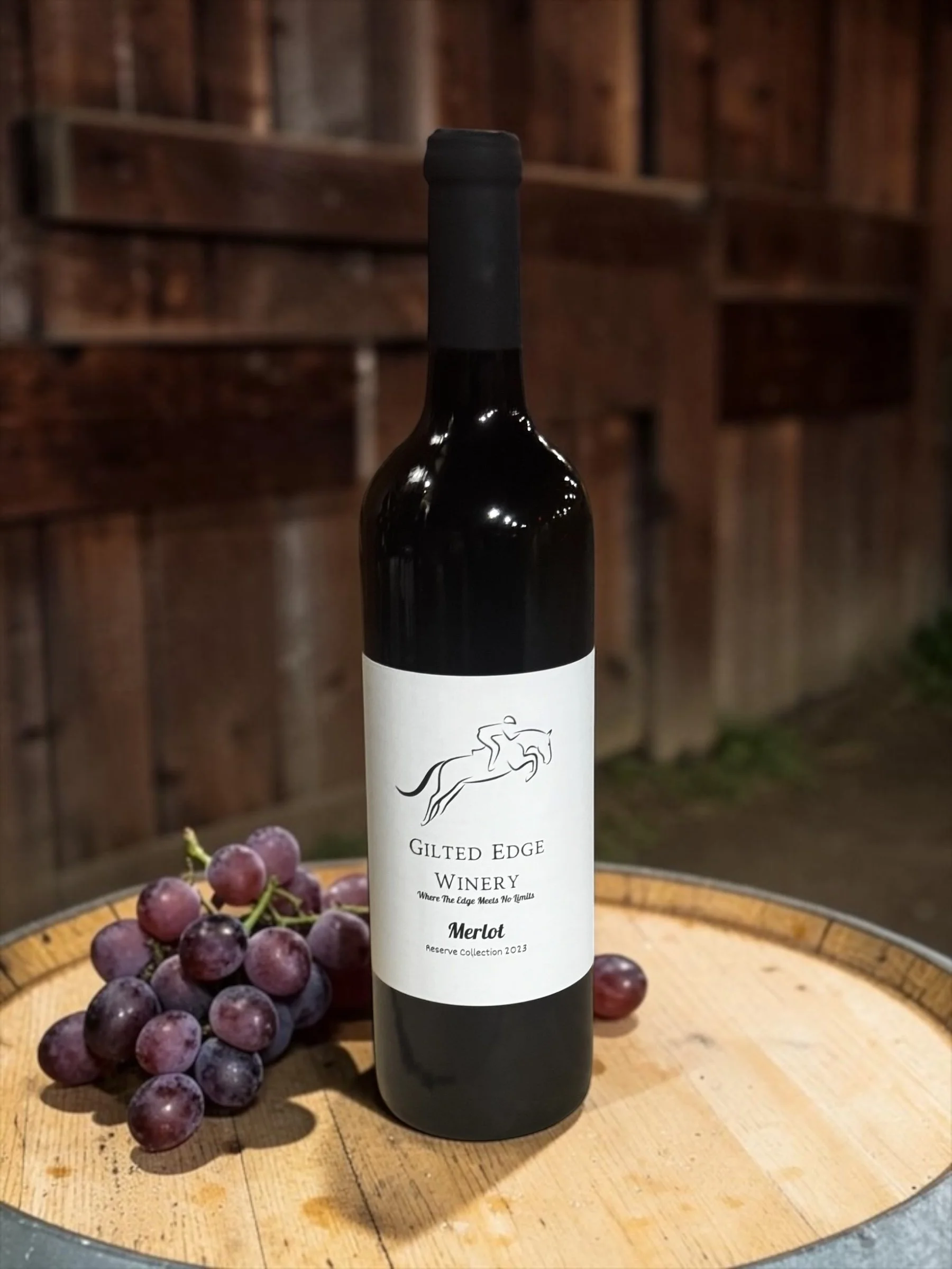 A bottle of Gilted Edge Winery Merlot wine placed on a wooden surface next to a bunch of purple grapes.