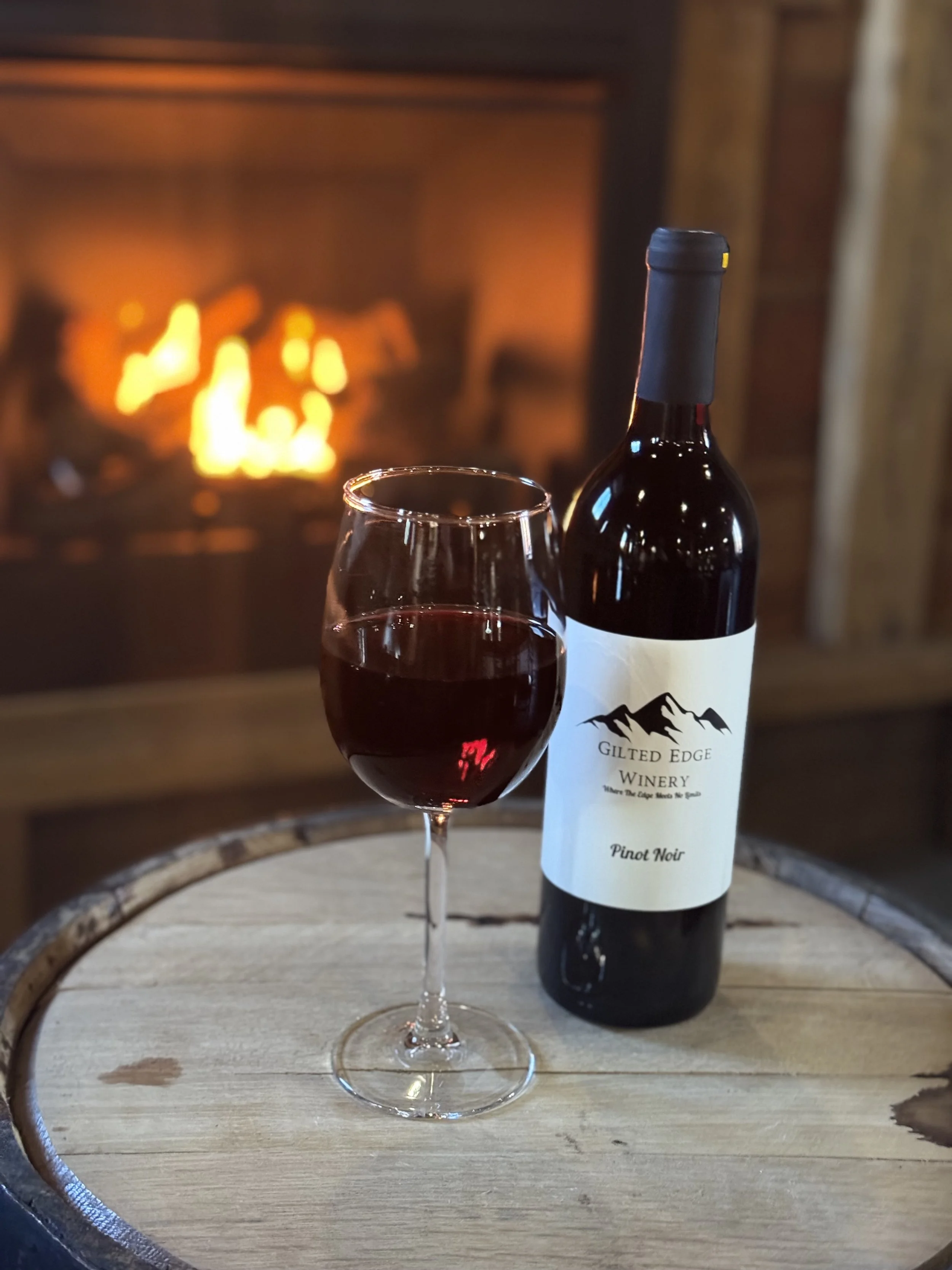 A glass of red wine and a bottle of Gilt Edge Winery Pinot Noir on a wooden barrel in front of a fireplace.