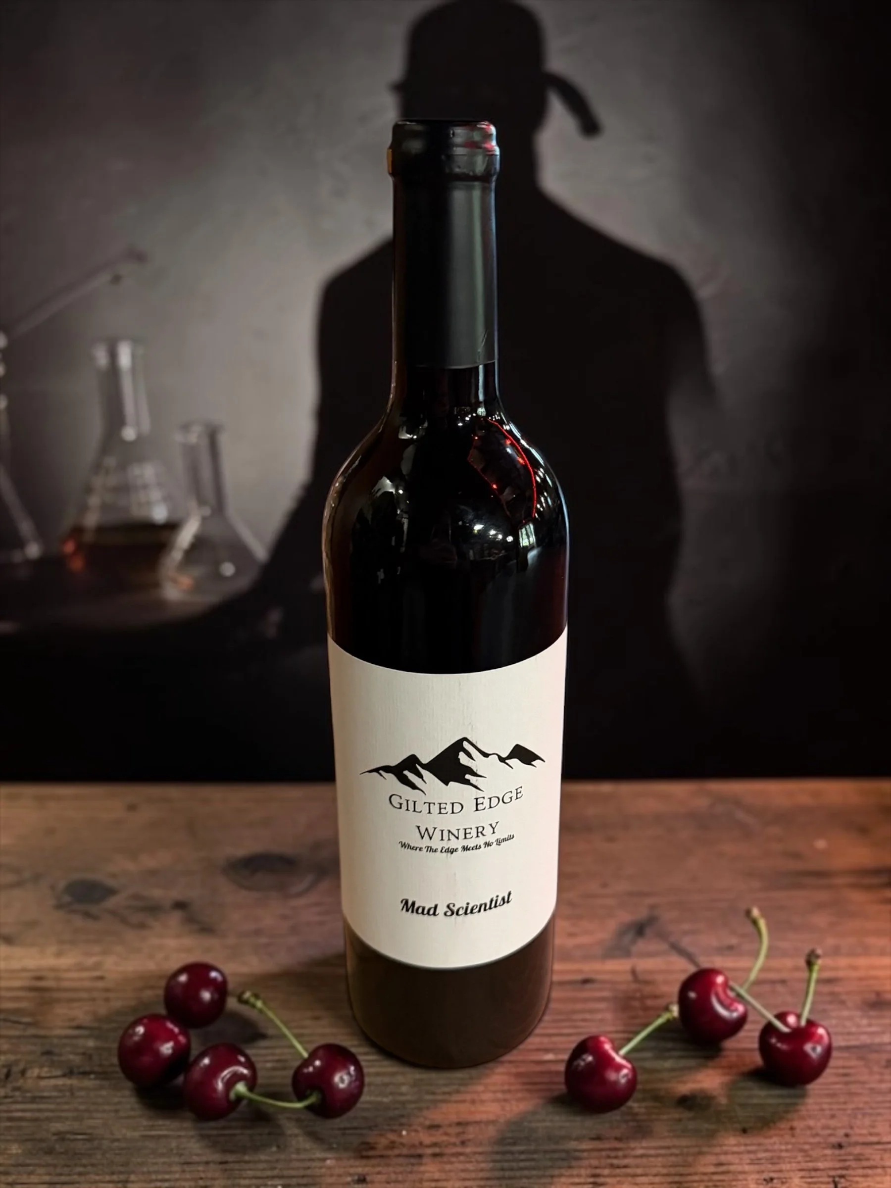 A wine bottle with a mountain logo and label reading Gilted Edge Winery and Mad Scientist, surrounded by cherries on a wooden surface. In the background, laboratory glassware is visible.