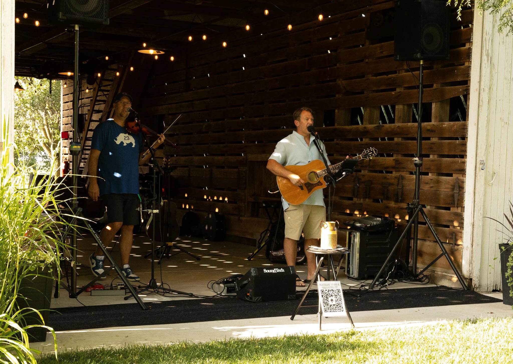 A live music performance on an outdoor stage with two musicians: one playing a violin and the other singing and playing an acoustic guitar. The stage has wooden paneling, string lights, and sound equipment, with greenery visible outside.