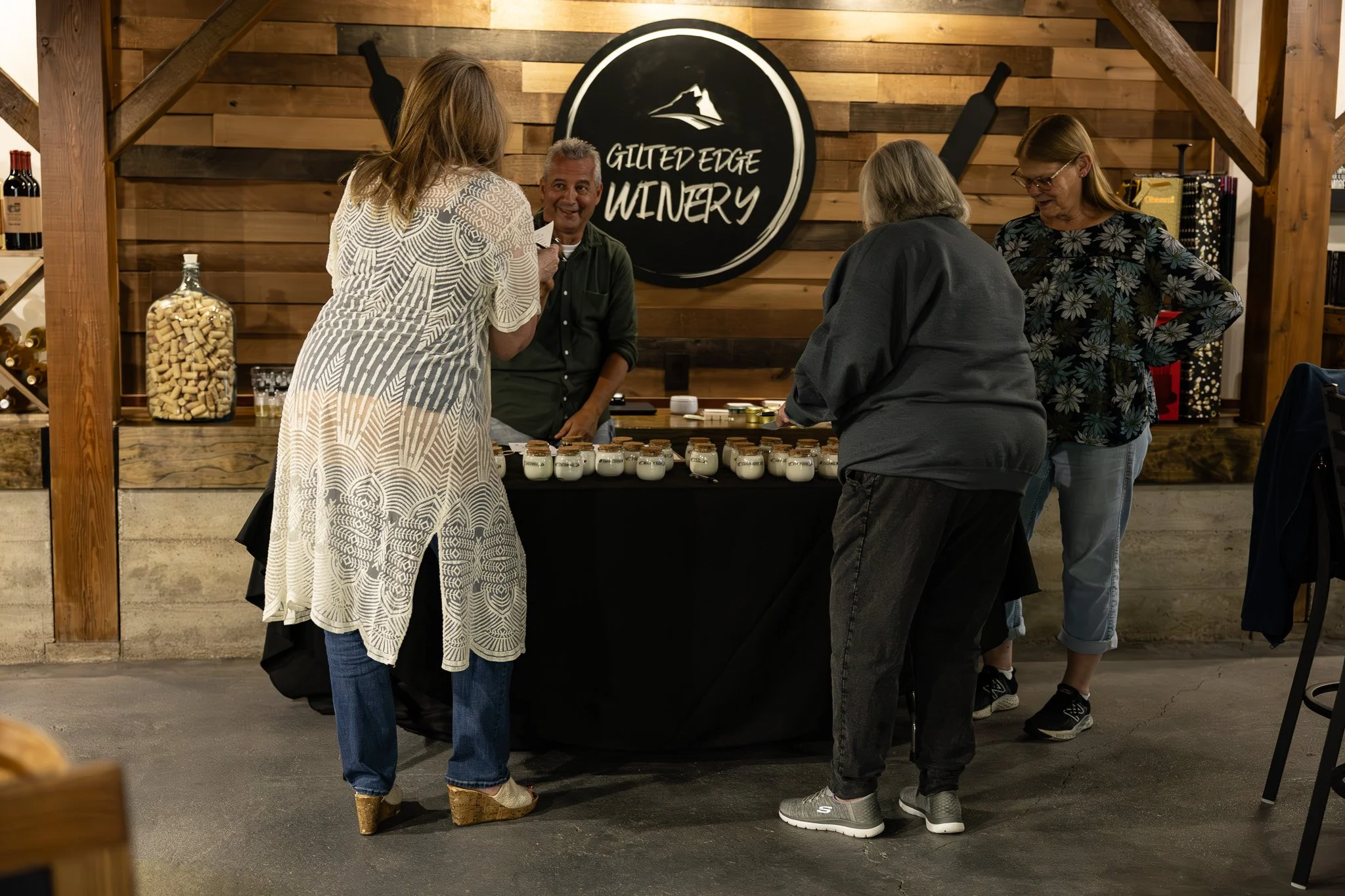 Four women and one man gathered around a table with small jars at Guiltedge Winery, with a wooden wall and winery logo in the background.