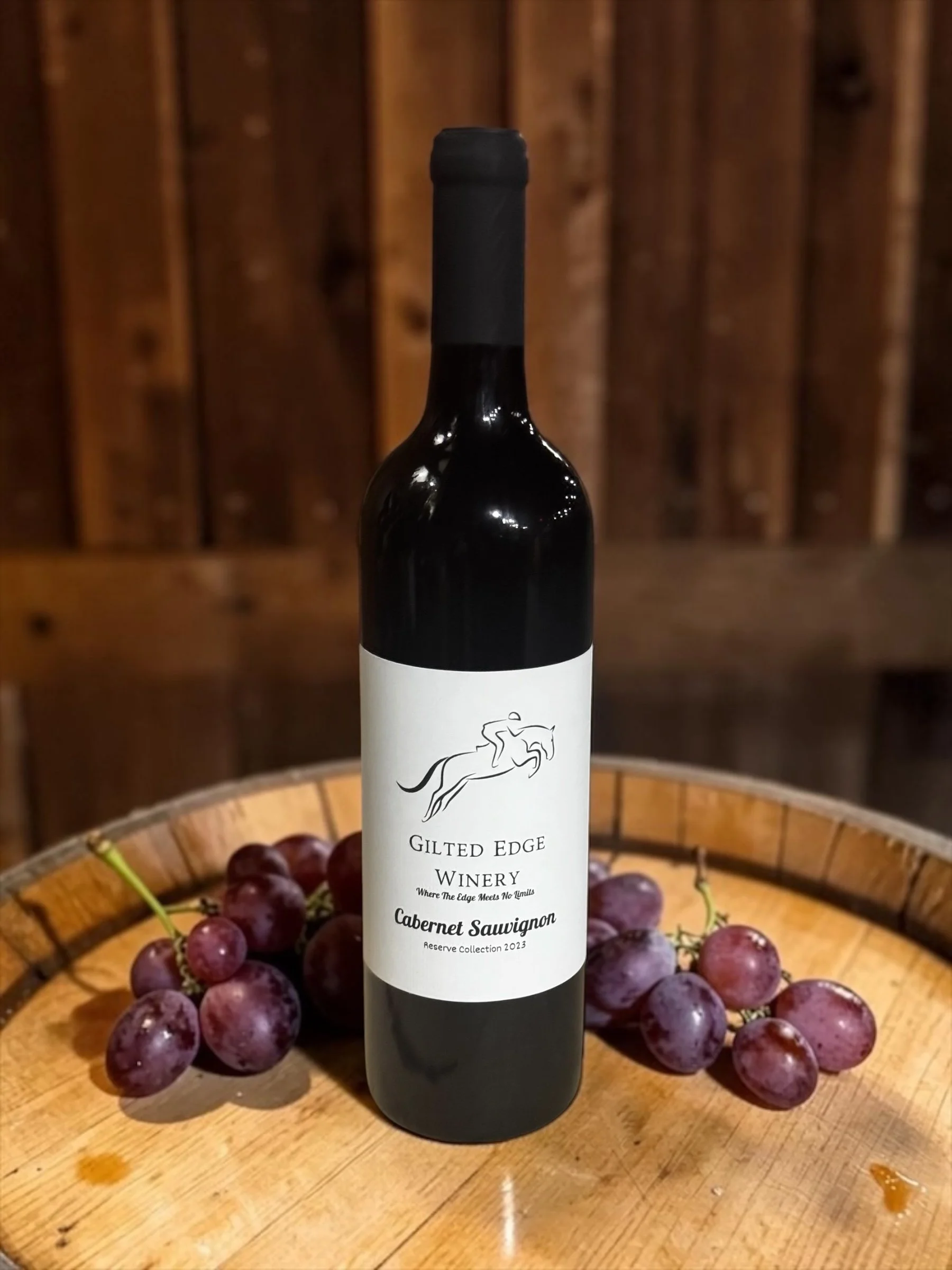 A bottle of Cabernet Sauvignon wine from Gilted Edge Winery placed on a wooden table or surface, surrounded by a bunch of purple grapes, with a wooden background.