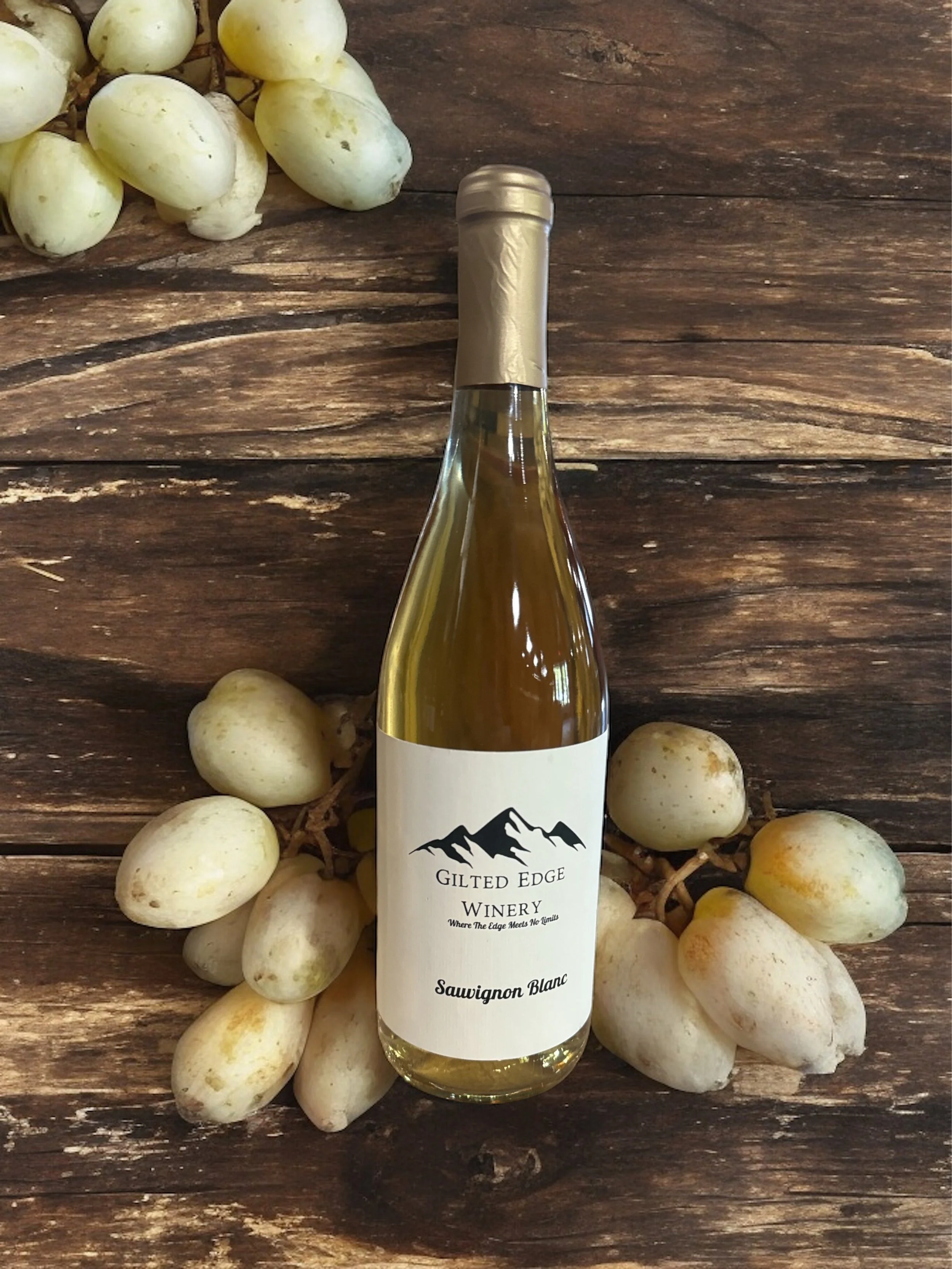 A bottle of wine from Gilded Edge Winery placed on a rustic wooden surface, surrounded by a bunch of white grapes.