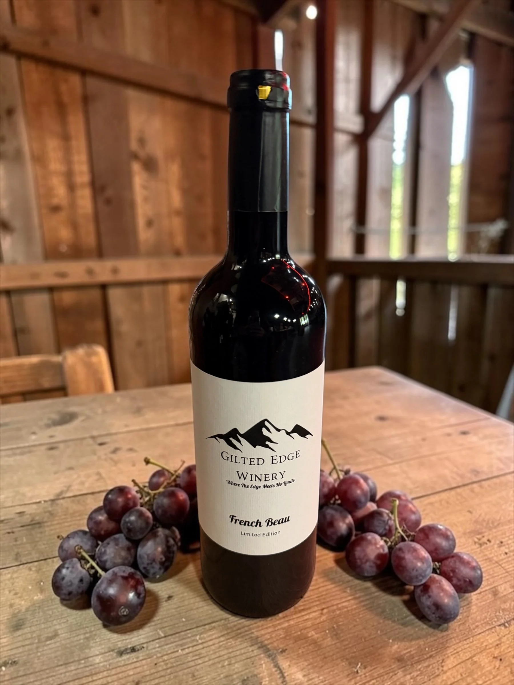 A bottle of Gilt Edge Winery wine titled French Beau with a mountain logo on the label, placed on a wooden table surrounded by red grapes, in a rustic wooden interior.