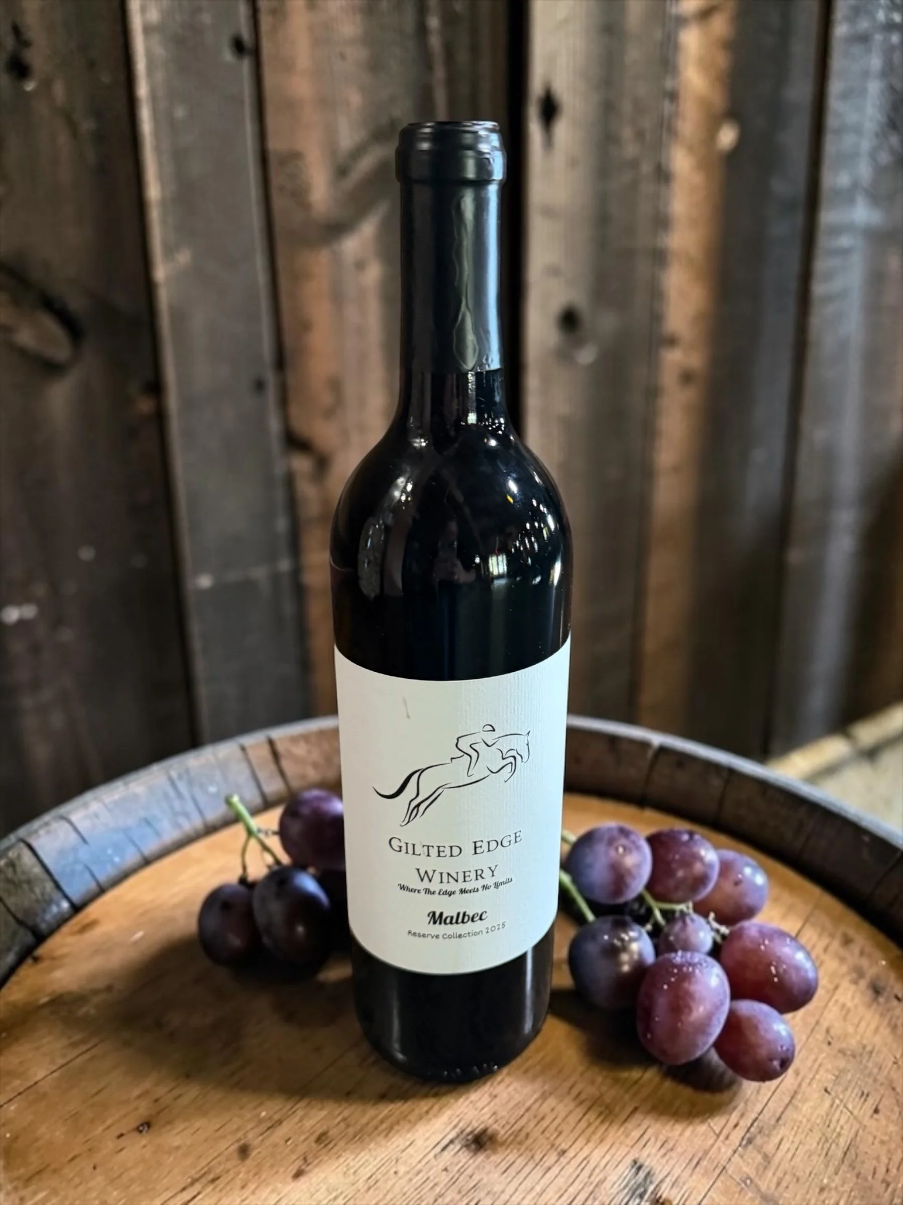 A bottle of Gilt Edge Winery Malbec wine surrounded by grapes on a wooden table with a rustic background.
