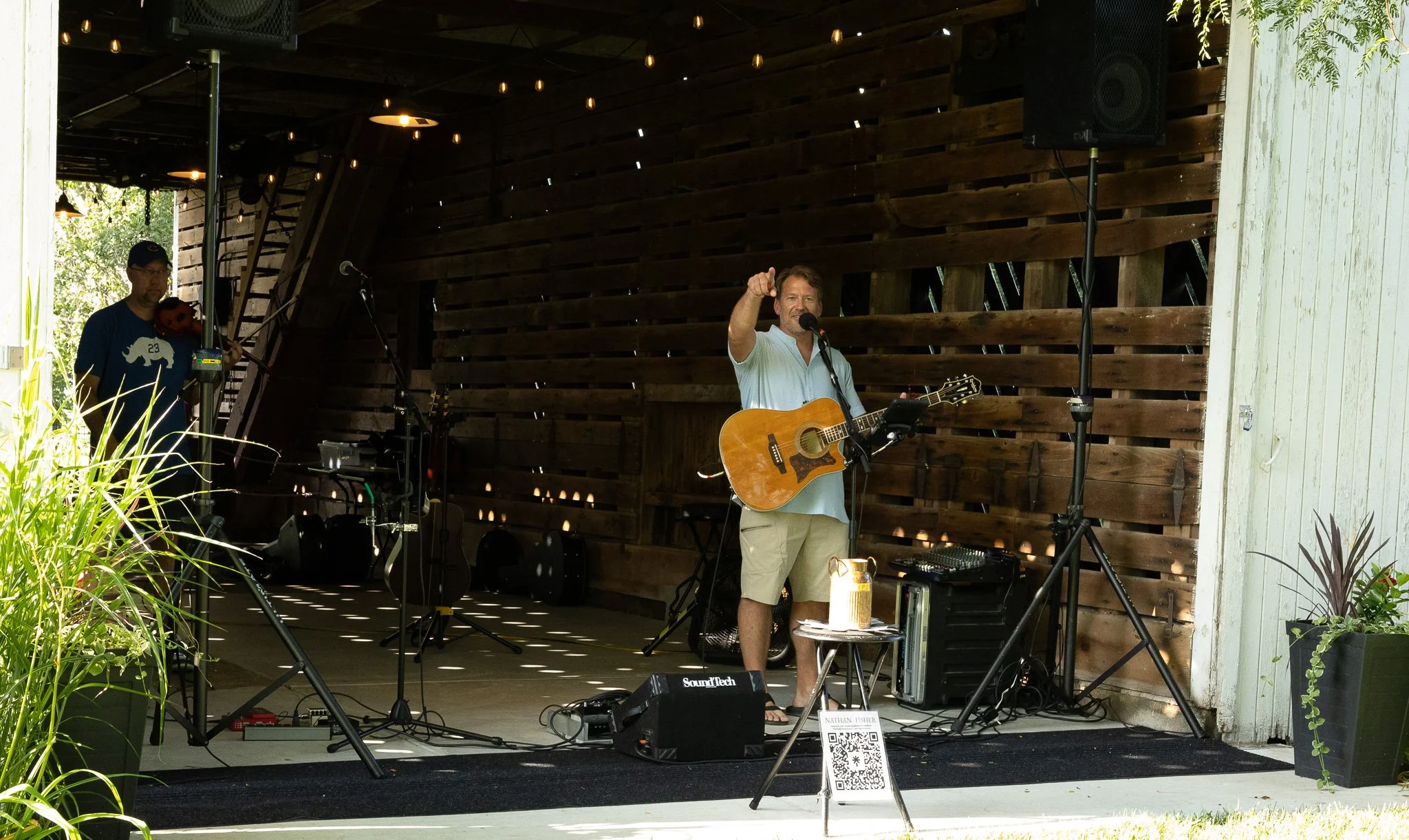 A man with a guitar singing into a microphone on a small stage, with another man playing a violin behind him. The environment appears to be a rustic, open-air venue with wooden walls and hanging lights.