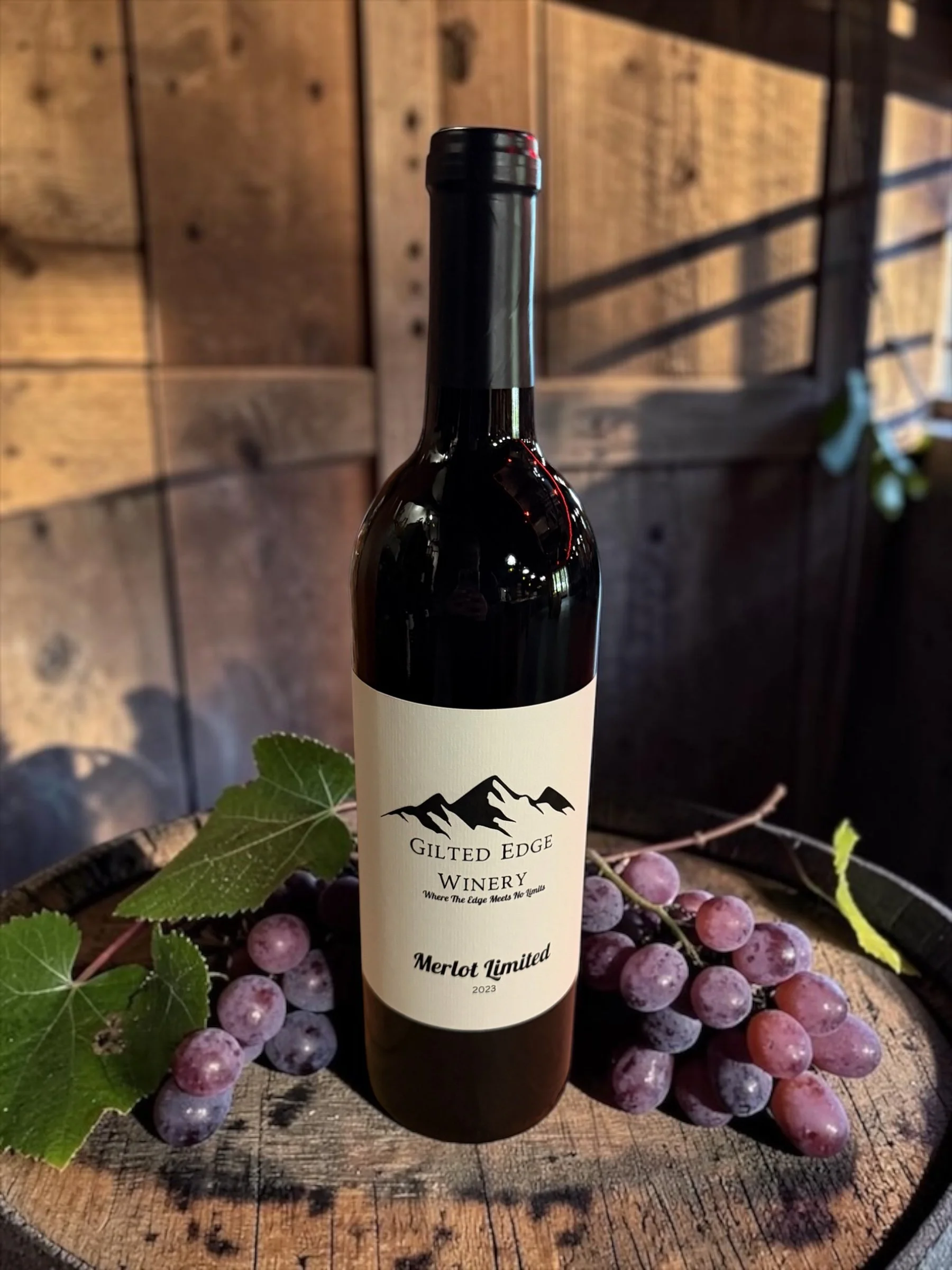A bottle of Gilt Edge Winery Merlot Limited wine surrounded by grapes and green leaves on a wooden surface.