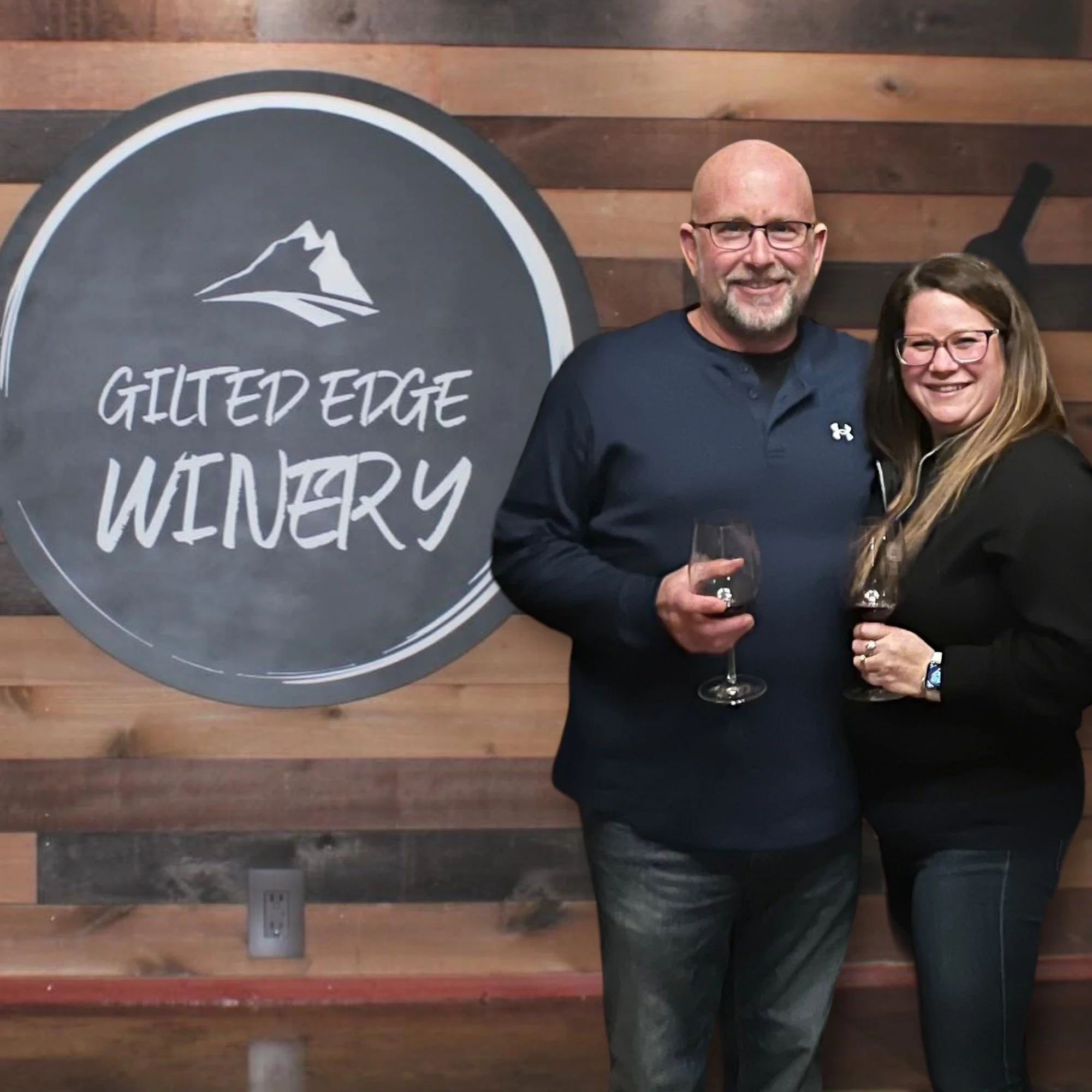 A man and a woman smiling and holding glasses of red wine in front of a wooden wall with a circular sign that reads 'Gilted Edge Winery' and features a mountain logo.