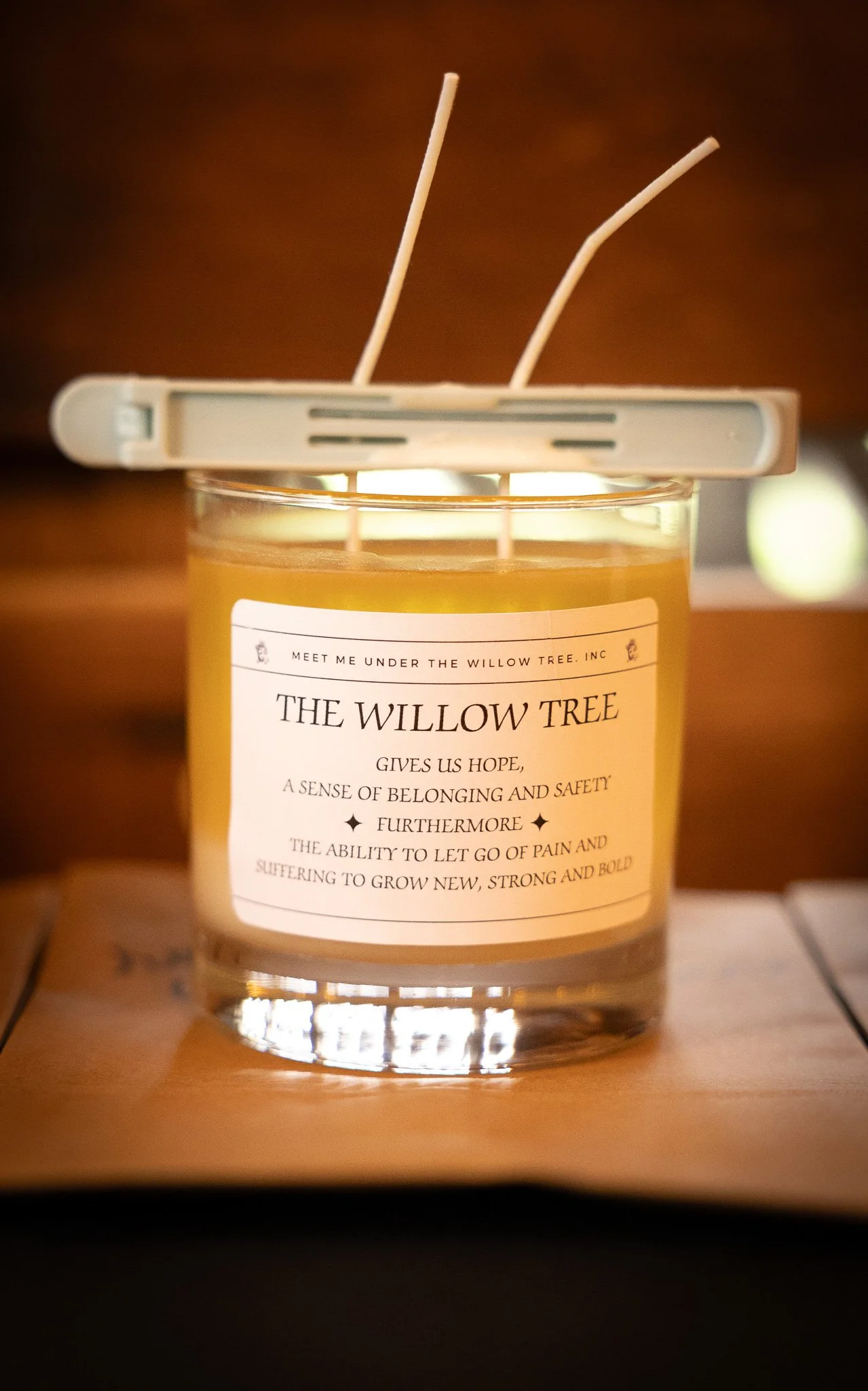 A lit candle in a glass jar with a label reading "The Willow Tree," with two wax sticks resting on top of the jar. The background is dark and blurred.