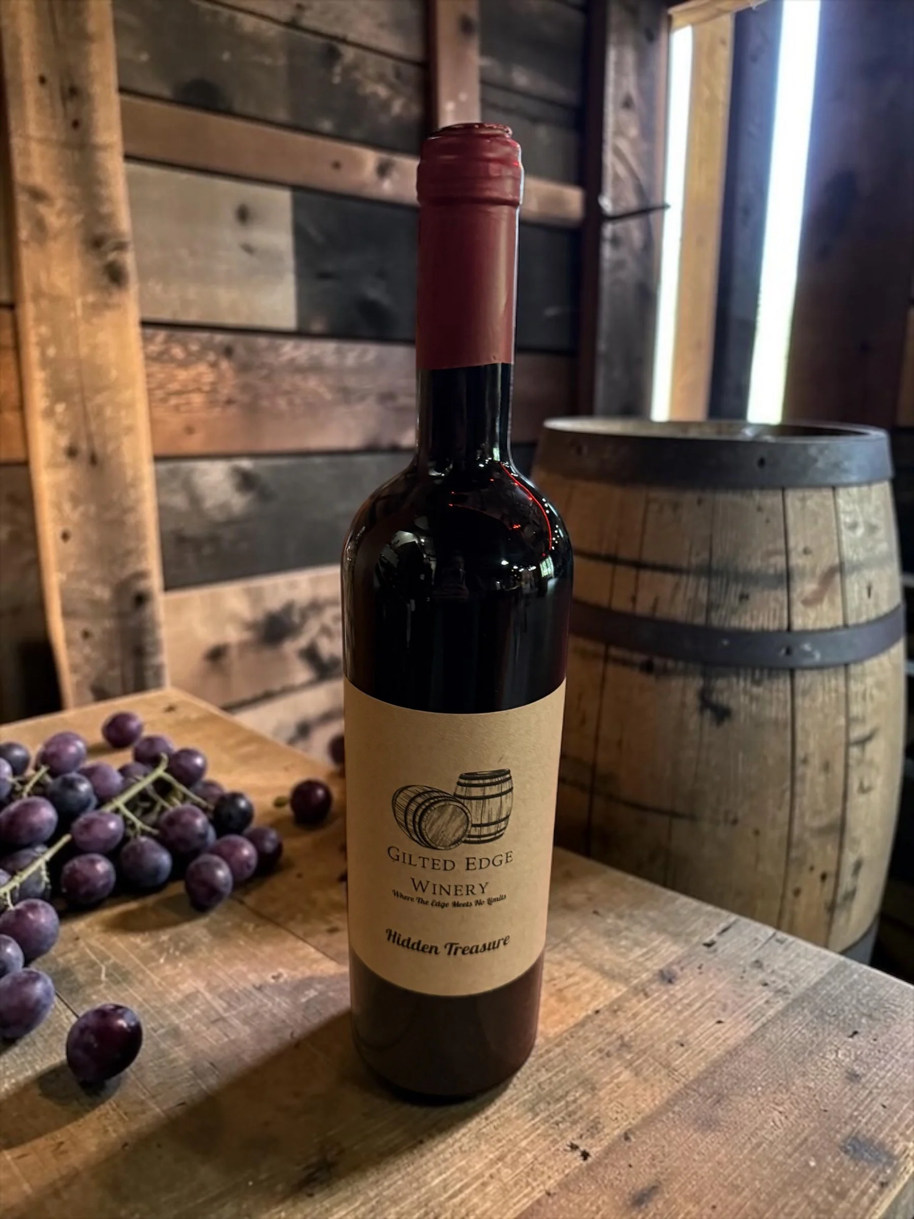 A wine bottle with a paper label that reads 'Gilded Edge Winery, Where the Edge Meets No Grains, Hidden Treasure' is placed on a wooden table. Nearby, there are grapes, and in the background, wooden barrels and a wooden wall are visible.