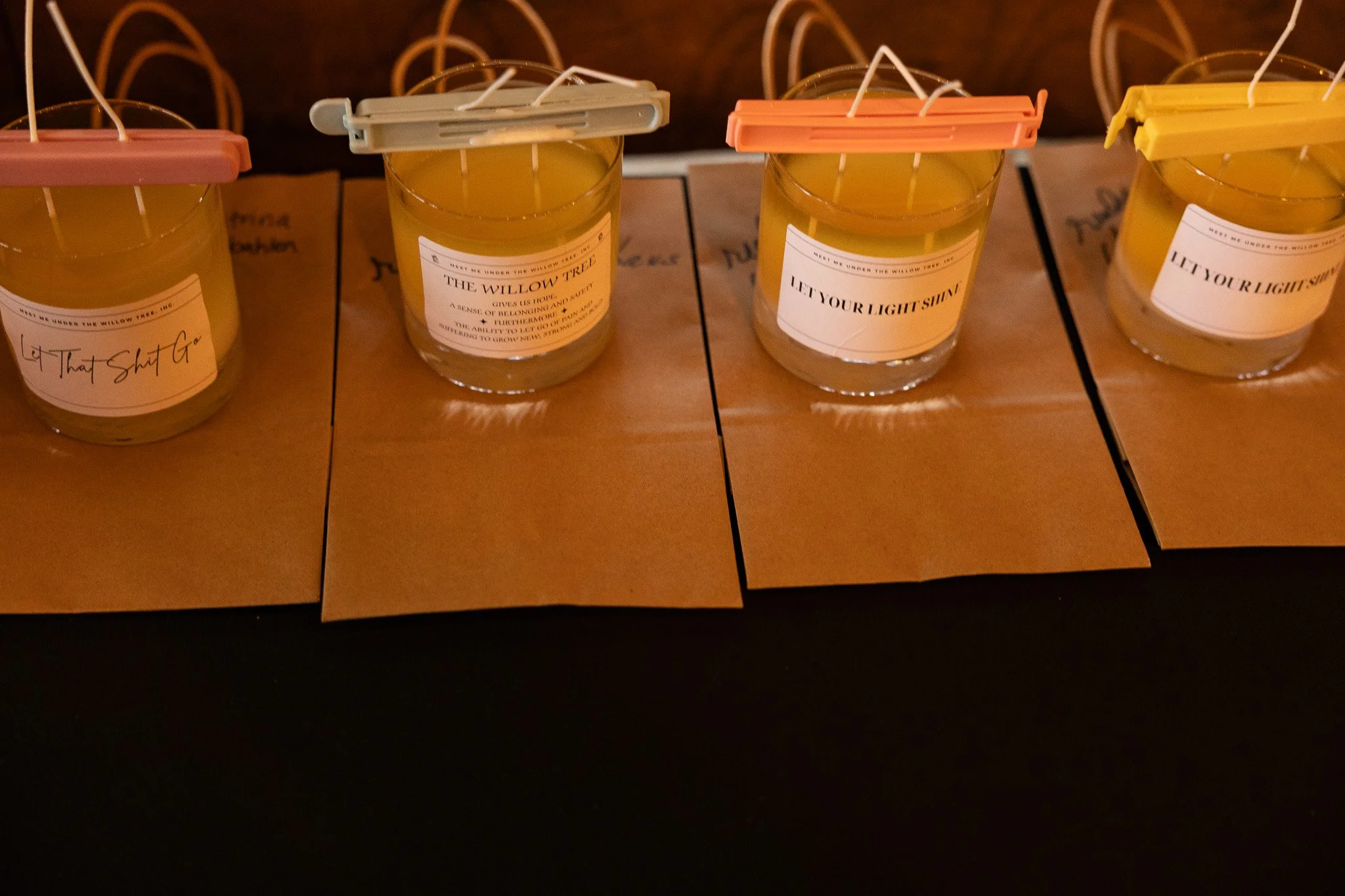 Four candles in glass jars on a wooden surface, each with a label featuring different messages. The first candle on the left has a label that says "That Shit G," and the remaining three have labels with various phrases: "THE WILLOW TREE," "LET YOUR L