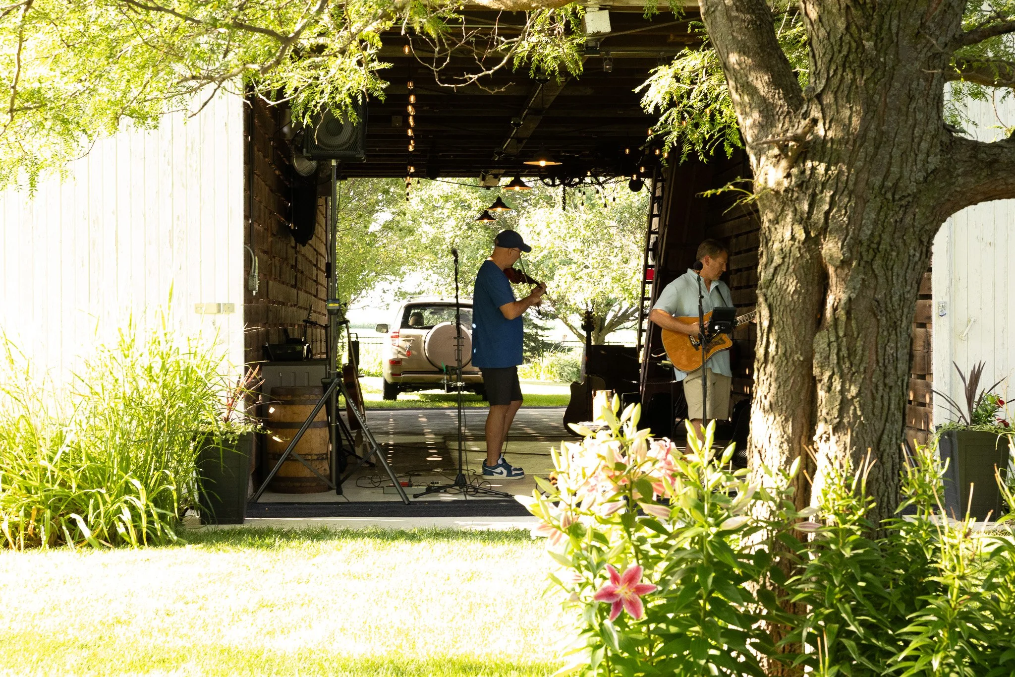 Two musicians perform on a small outdoor stage shaded by trees. One plays a guitar and sings into a microphone, while the other plays a violin.