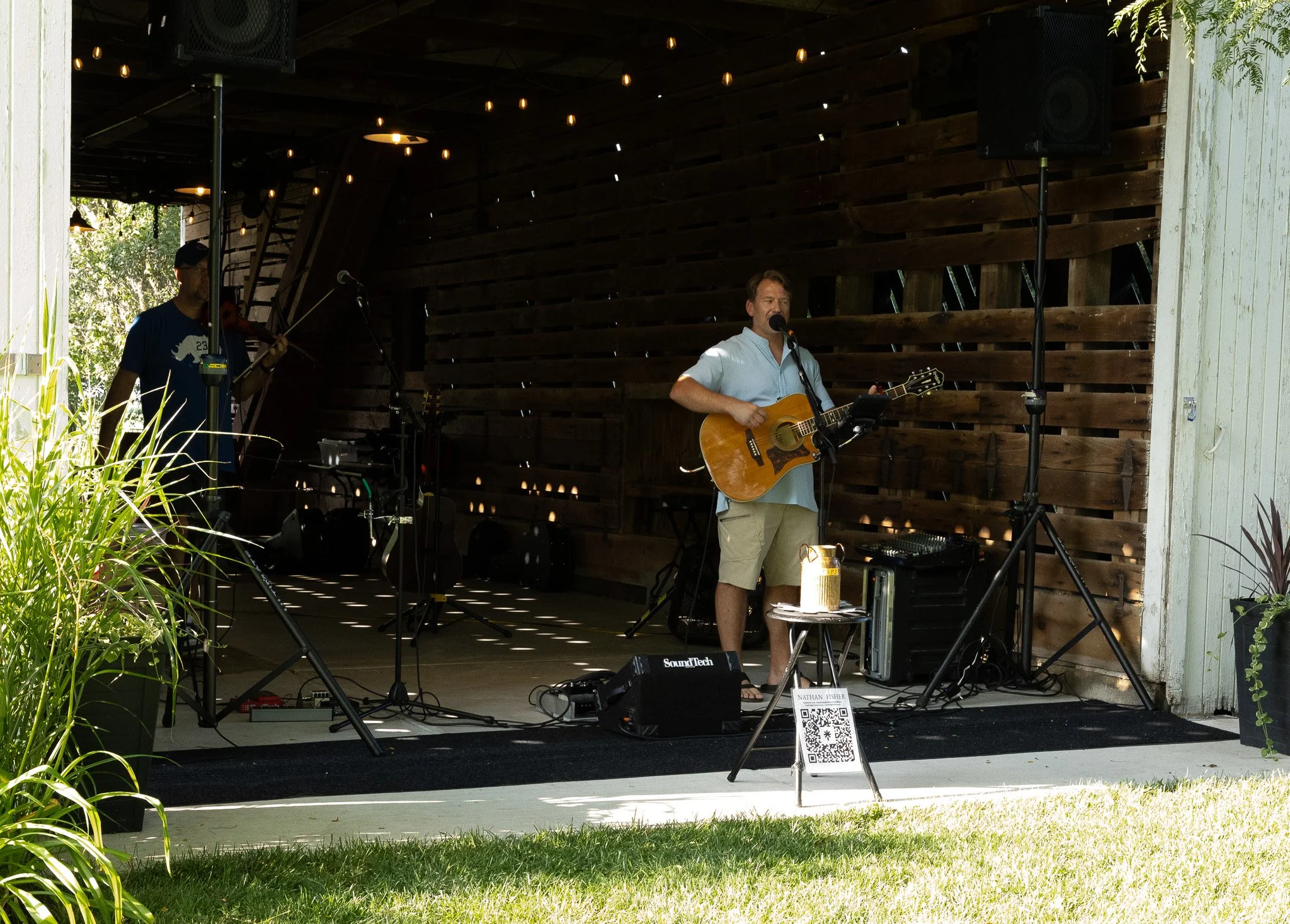 Male musician playing guitar and singing into a microphone on an outdoor stage with wooden walls and string lights, accompanied by a violinist in the background, with plants and foliage around the stage.