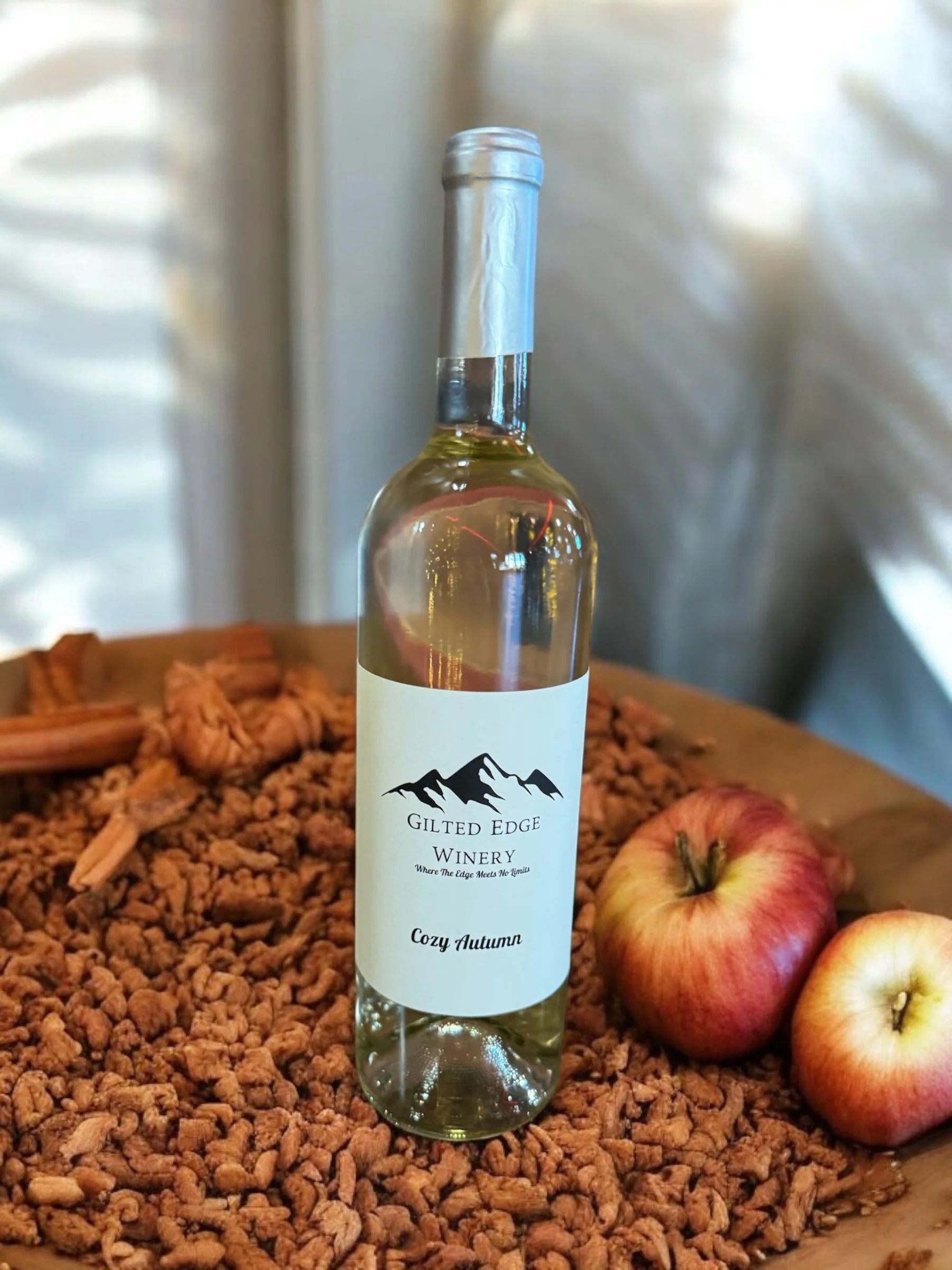 A clear bottle of wine labeled 'Gilted Edge Winery Cozy Autumn' stands on a bed of small brown wood chips, alongside three red and yellow apples, with a blurred background.