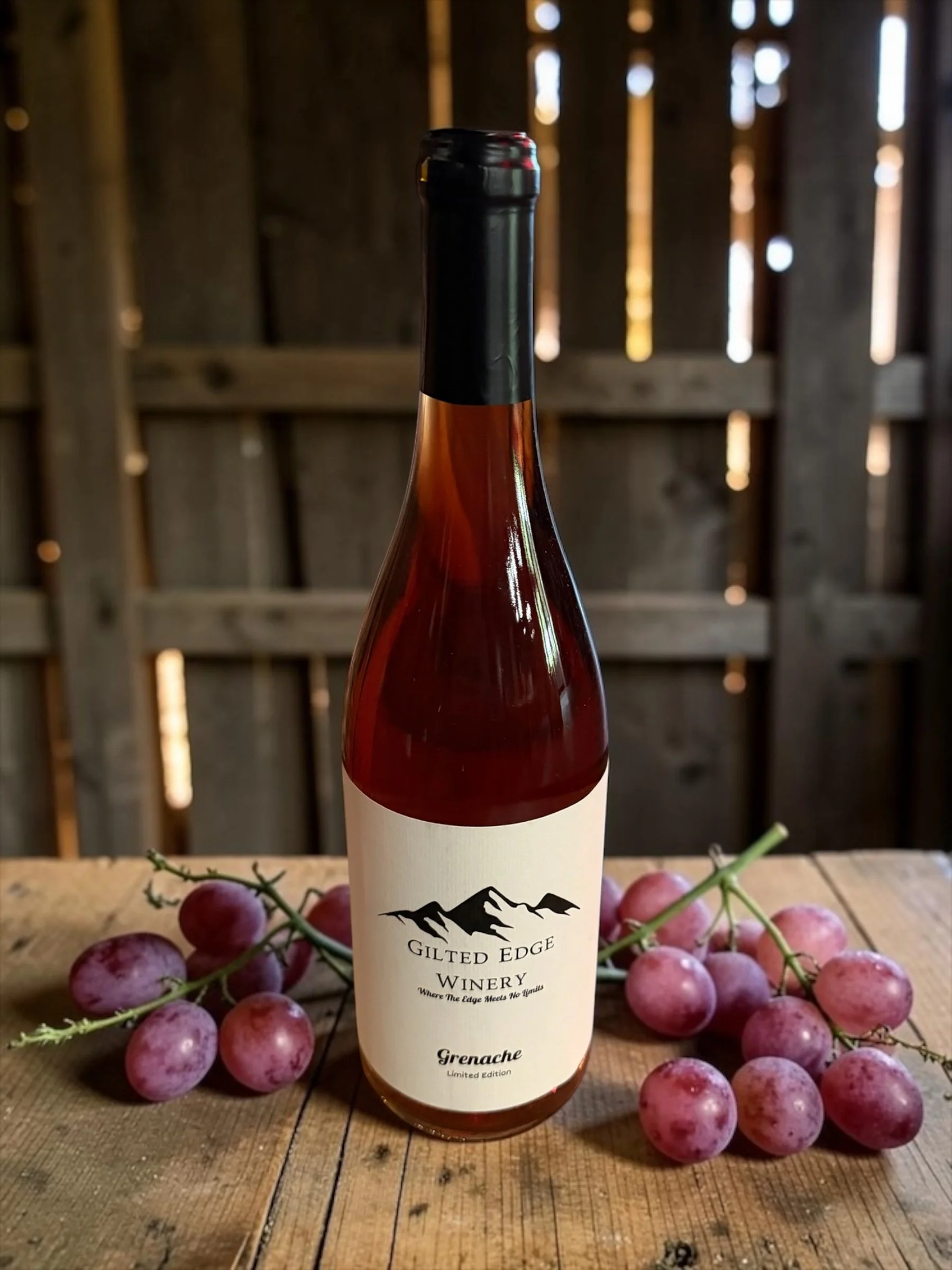 A bottle of Grenache wine from Gilt Edge Winery surrounded by purple grapes on a wooden surface, in a rustic setting with wooden walls in the background.