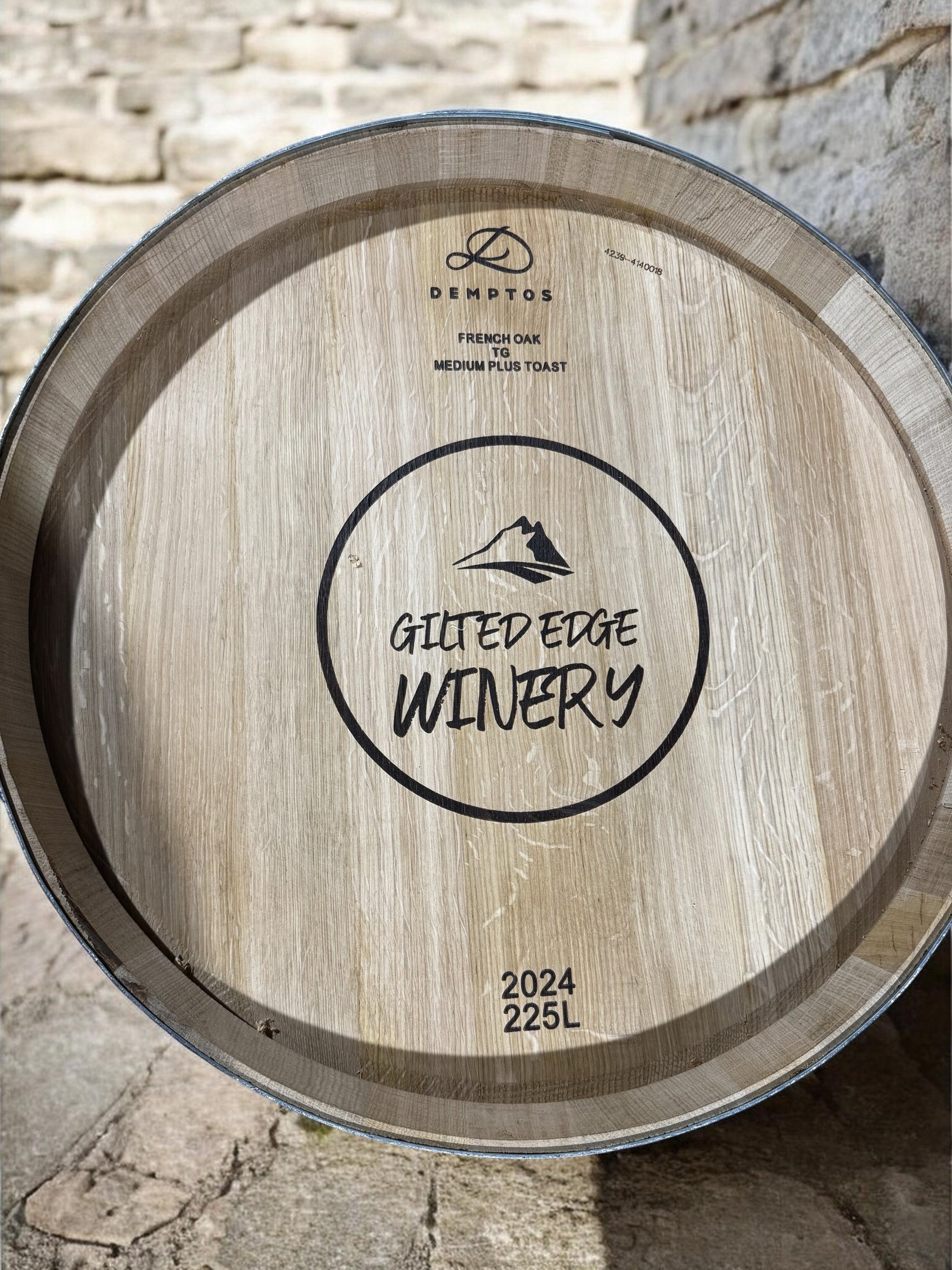 Close-up of the bottom of a wine barrel with labeled information including winery name, vintage year 2024, and capacity of 225 liters, against a stone wall background.