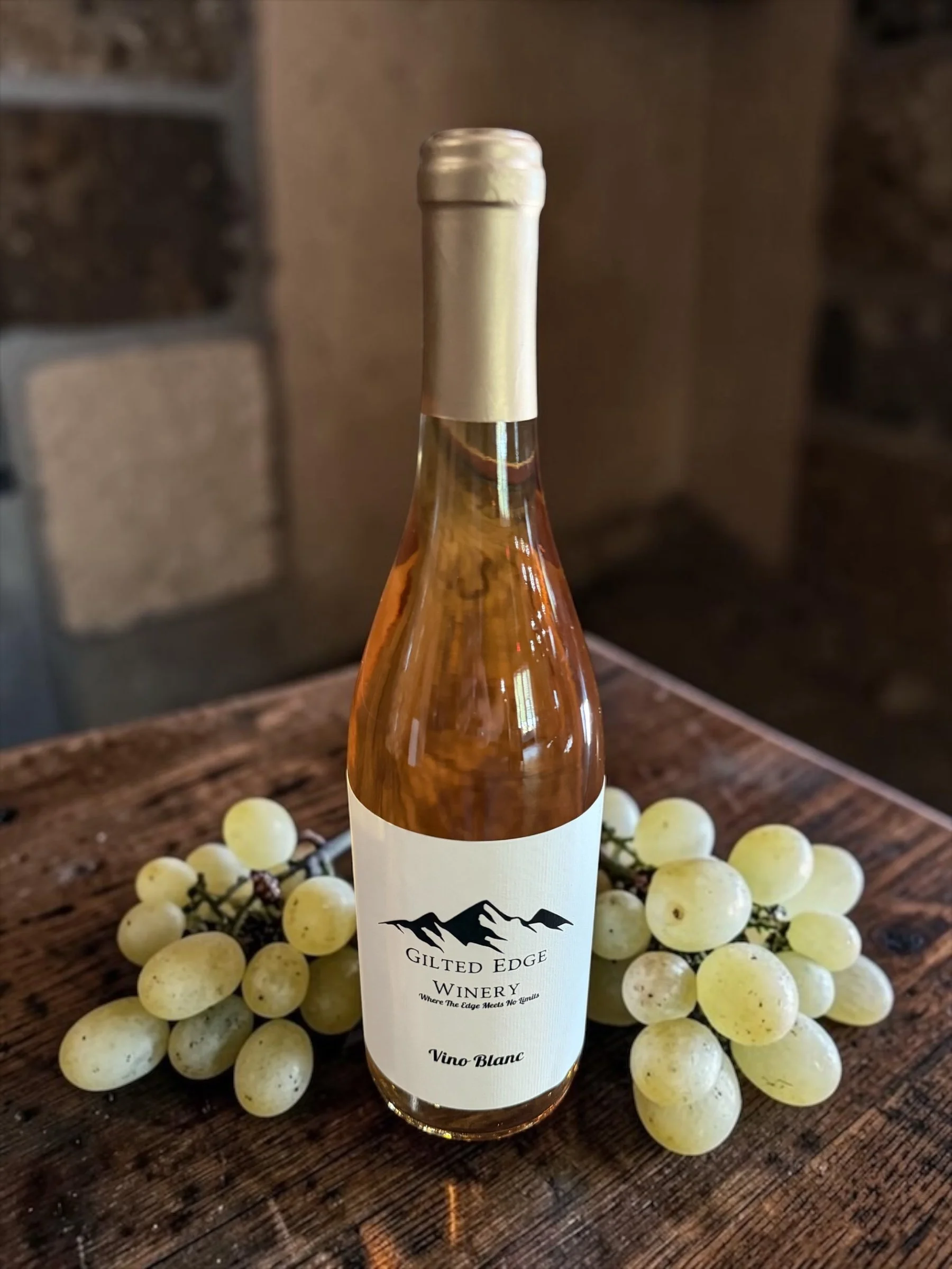 A bottle of white wine labeled 'Gilt Edge Winery' with a mountain logo, placed on a rustic wooden surface surrounded by a bunch of green grapes.