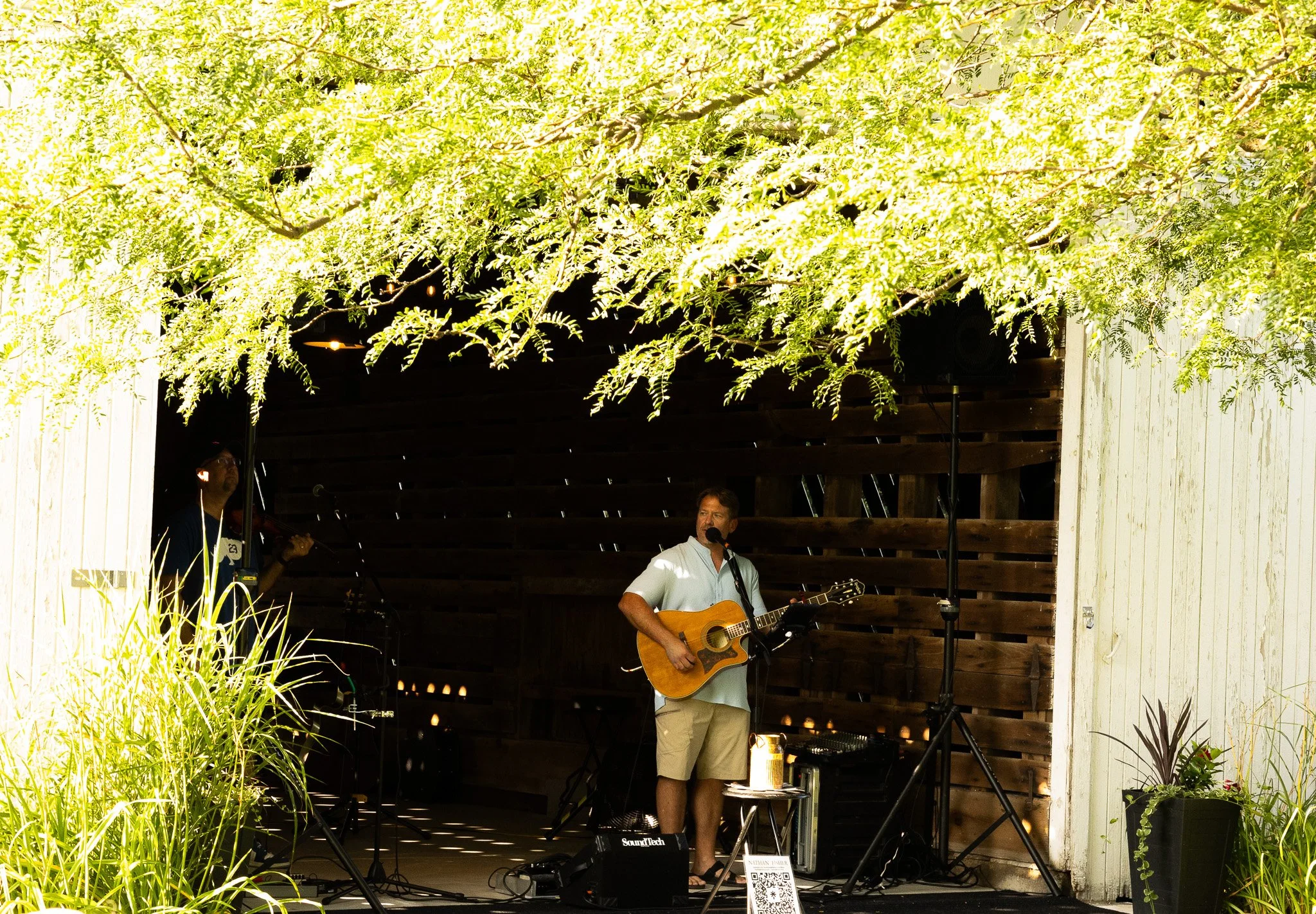 A man playing an acoustic guitar and singing into a microphone on an outdoor stage with greenery and plants around.