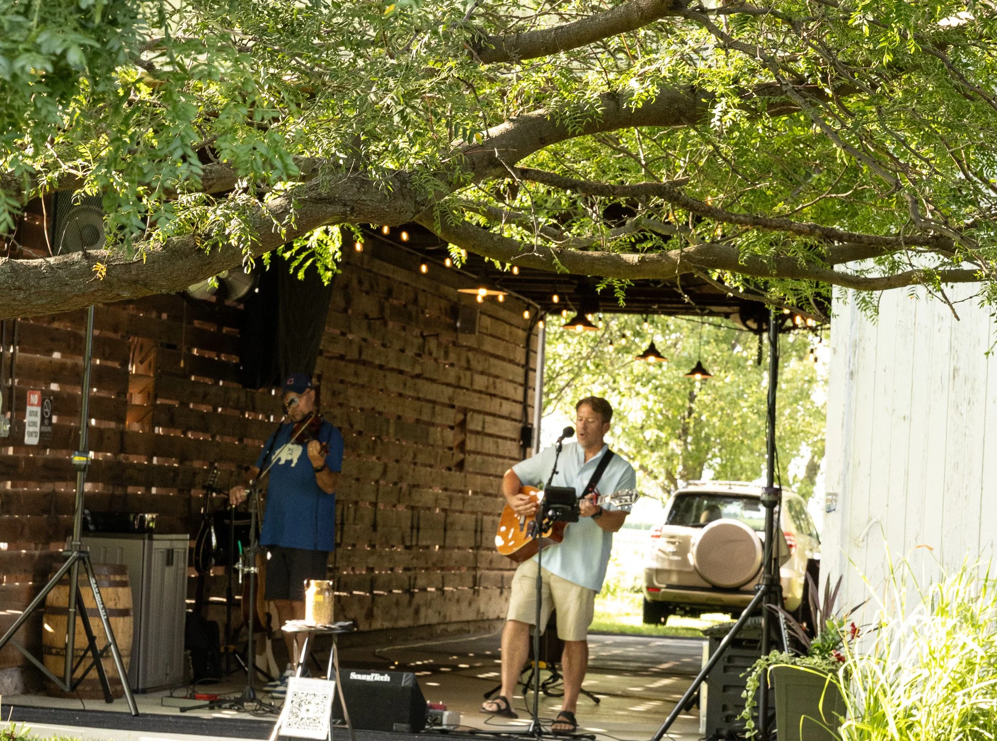 Two musicians perform outdoors under string lights; one plays a guitar, the other sings into a microphone, with a wooden fence and trees in the background.