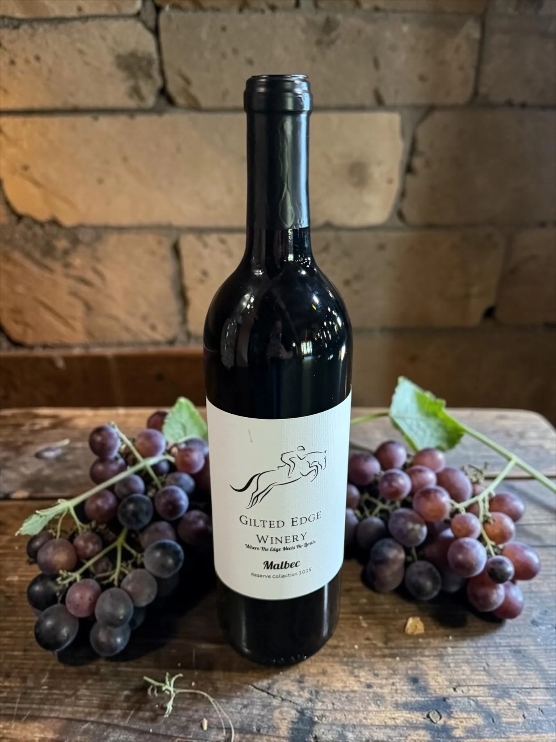 A bottle of Gilded Edge Winery Malbec wine with a minimalistic label featuring a stylized leaping animal and wine description, placed on a rustic wooden table surrounded by bunches of red and black grapes, with a background of a brick wall.