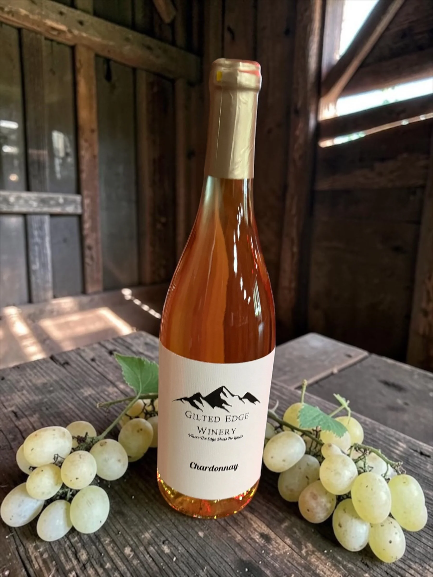 A bottle of Chardonnay wine from Gilt Edge Winery, surrounded by green grapes on a rustic wooden surface with grape leaves.