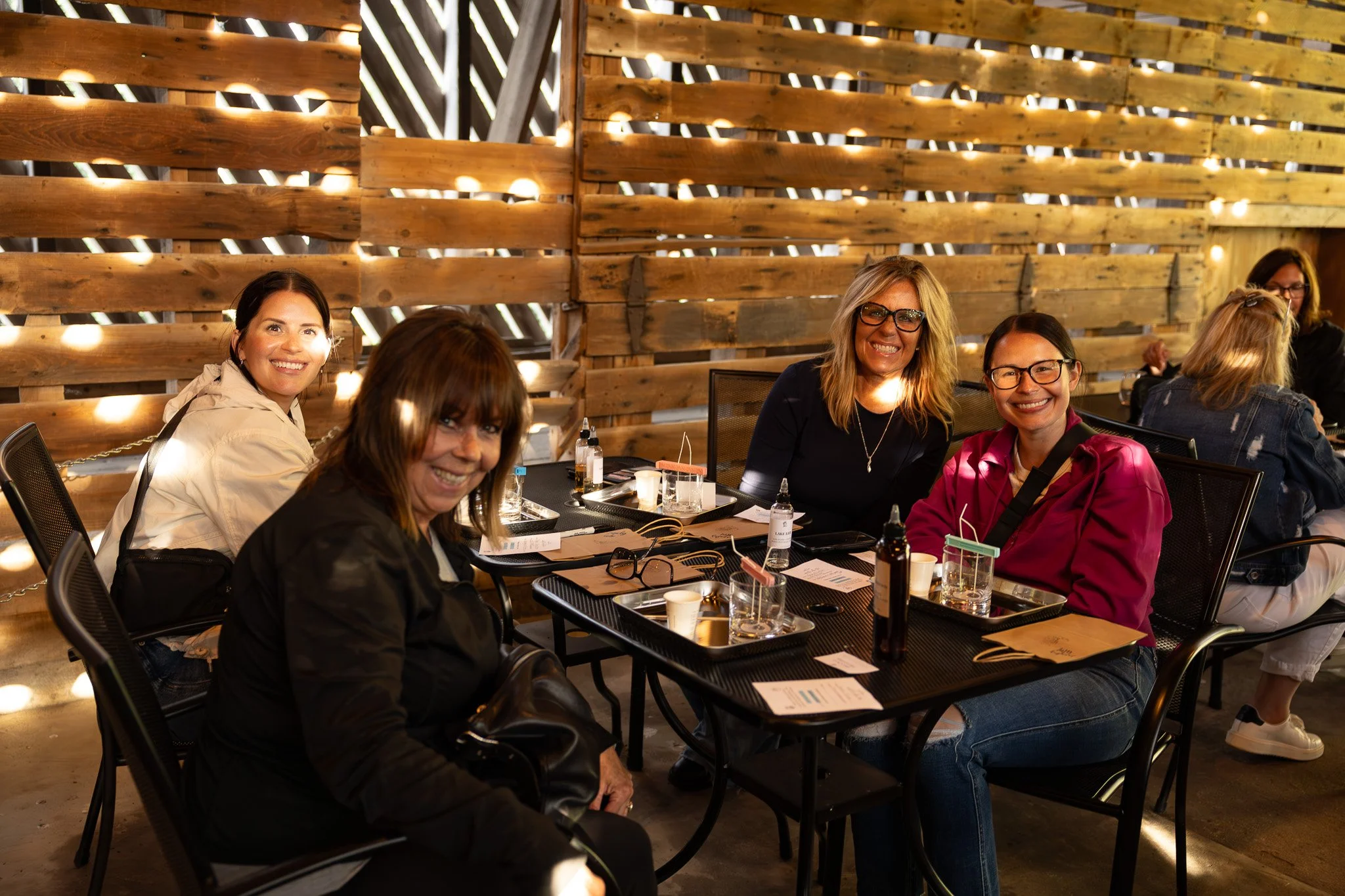 A group of women sitting at a table, smiling and enjoying a gathering in a rustic wooden venue with wooden plank walls and sunlight filtering through gaps.