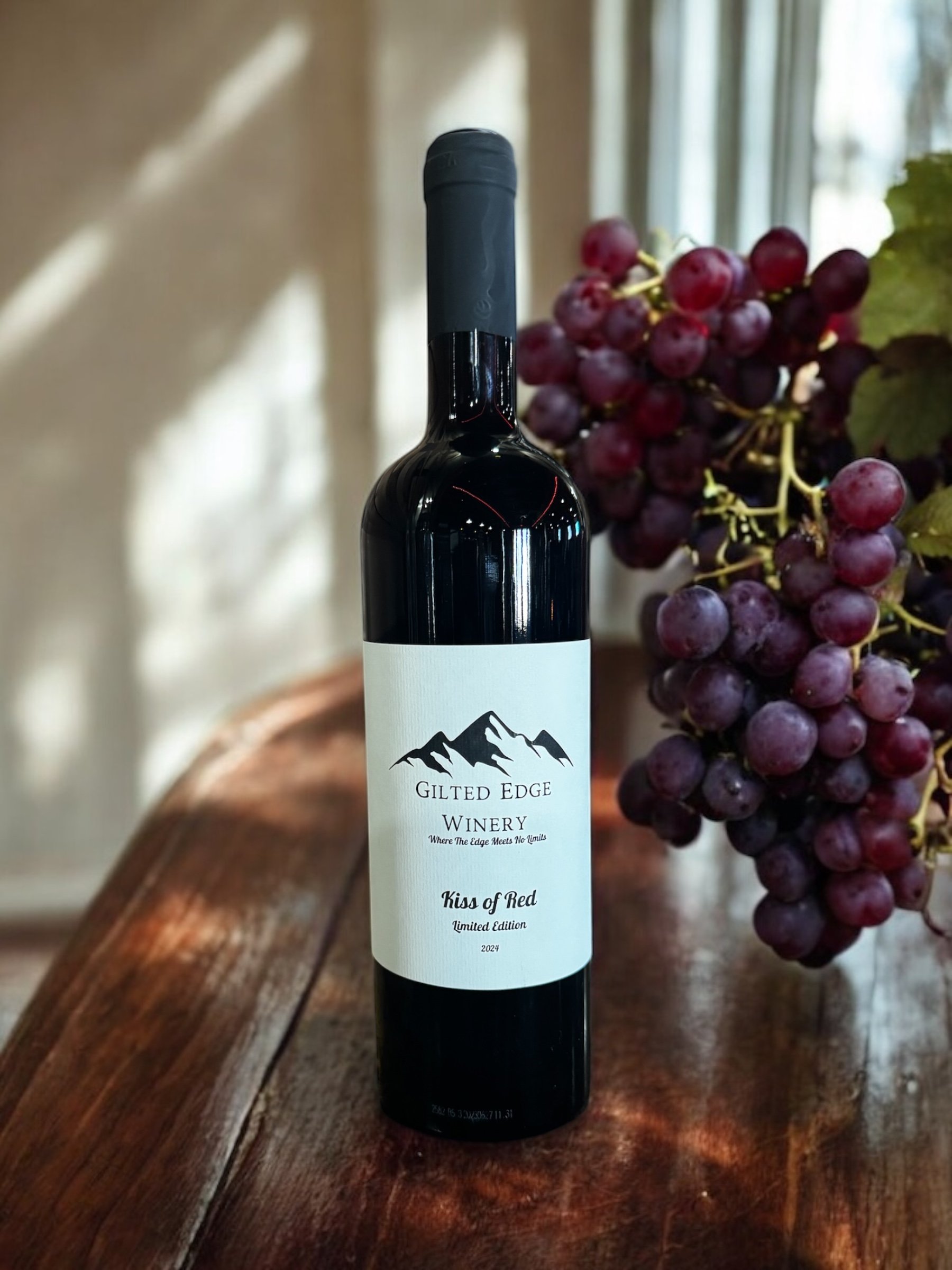 A bottle of red wine labeled 'Kiss of Red,' from Gilt Edge Winery, placed on a wooden surface with a bunch of red grapes next to it and a background of a window with curtains and a potted plant.