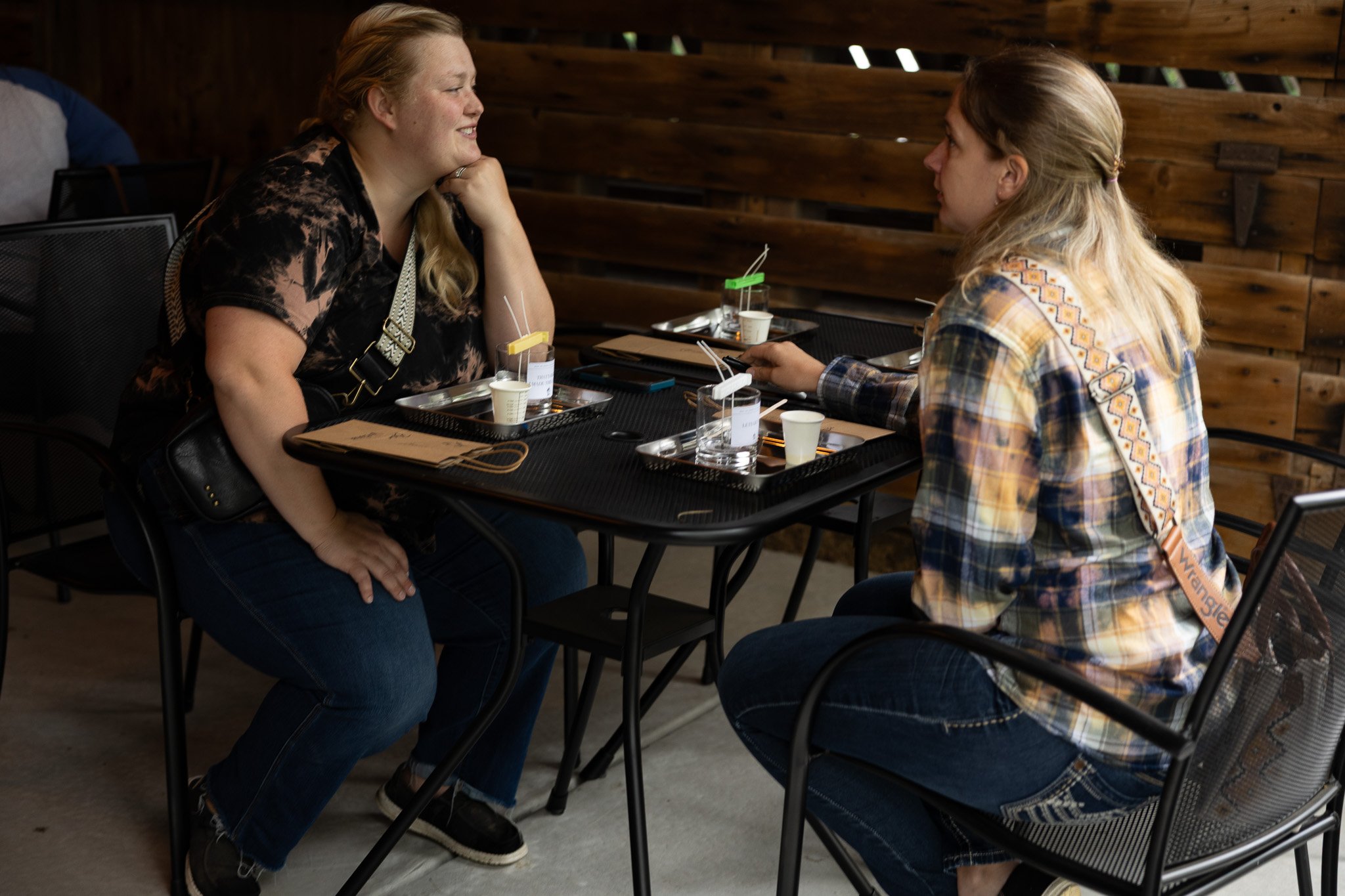 Two women sitting at a table in a cozy, wood-paneled restaurant, engaging in conversation. One woman has long, blonde hair and is wearing a plaid shirt, while the other has long, reddish hair and is wearing a dark patterned top.