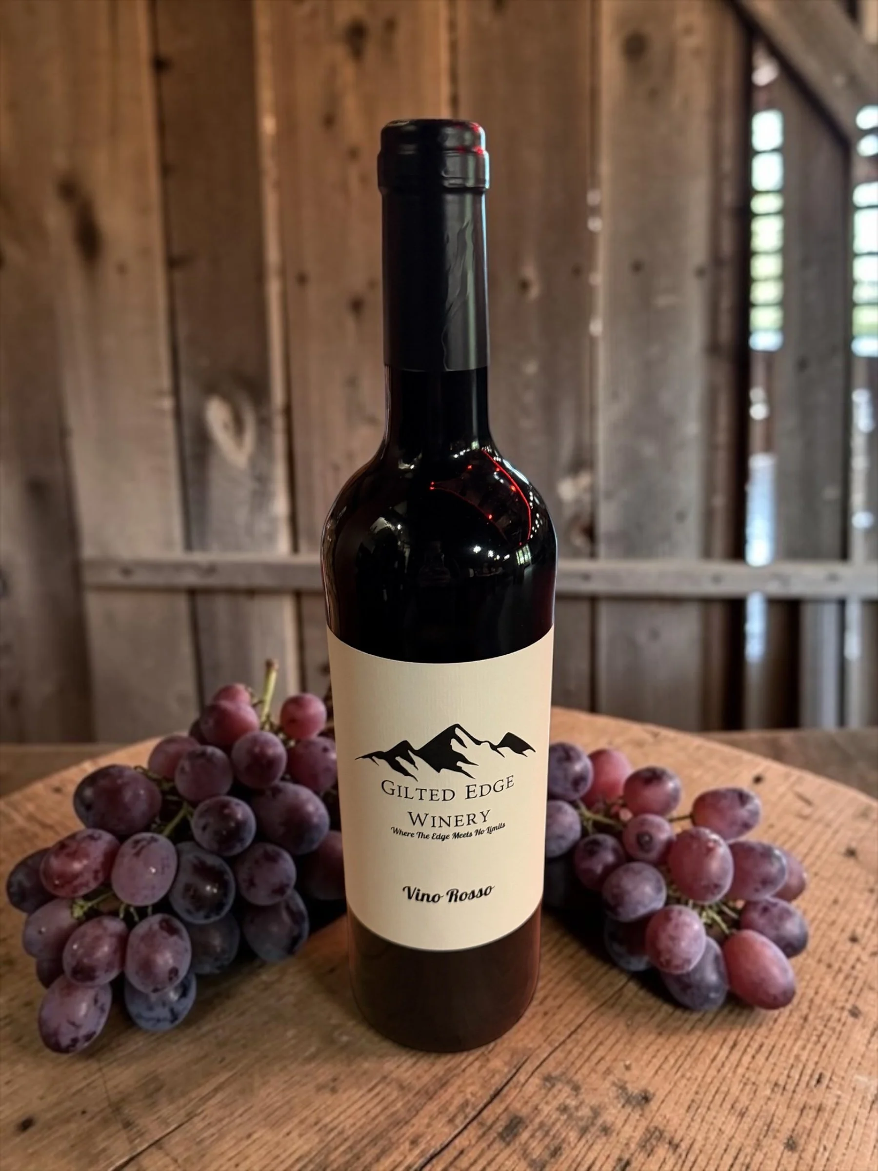 A bottle of wine labeled Gilded Edge Winery with grapes around it on a wooden table, set against a rustic wooden background.
