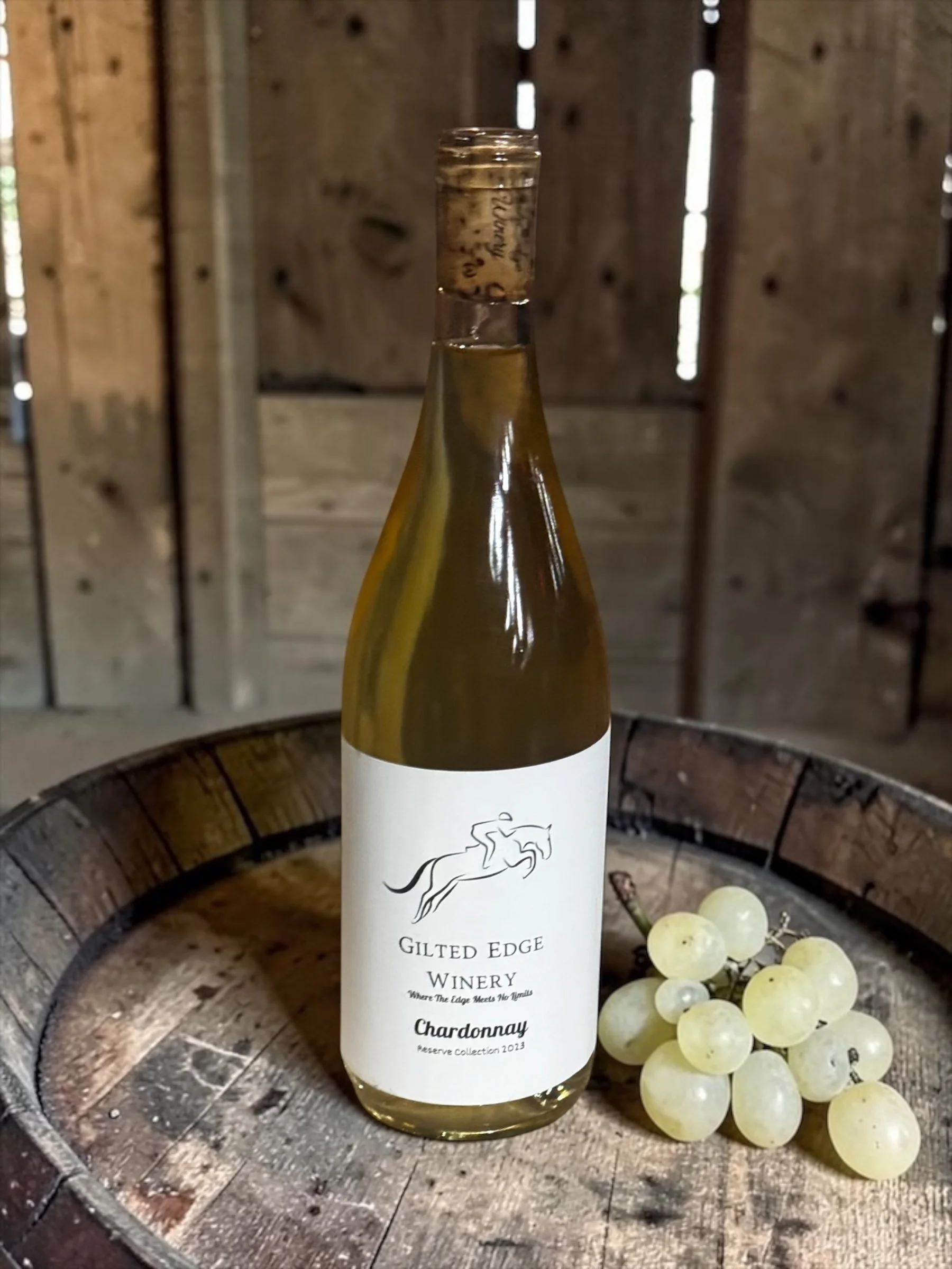 A bottle of white wine labeled 'Gilted Edge Winery, Chardonnay, Reserve Collection 2023,' placed on a rustic wooden surface with a cluster of green grapes beside it.