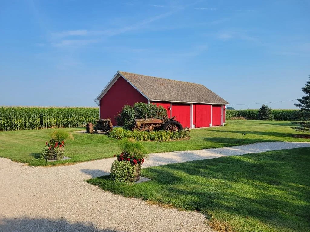 A red barn with a gray roof sitting on a lush green lawn, with an old tractor in front, surrounded by flowerpots and a cultivated cornfield in the background under a blue sky. Original Red Barn on site.