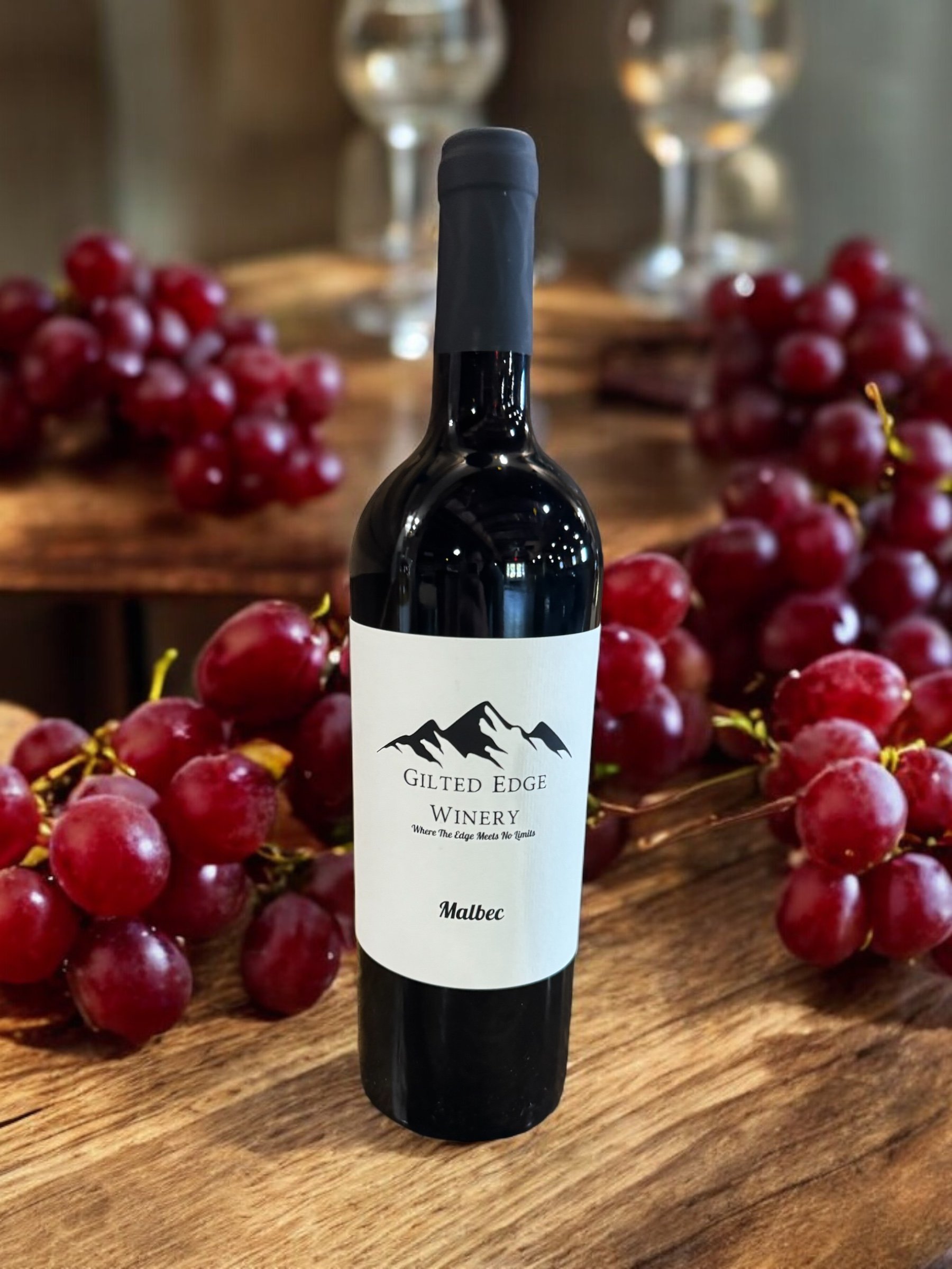 A bottle of Malbec wine from Gilded Edge Winery is placed on a wooden surface, surrounded by bunches of red grapes, with a blurred background of glassware.