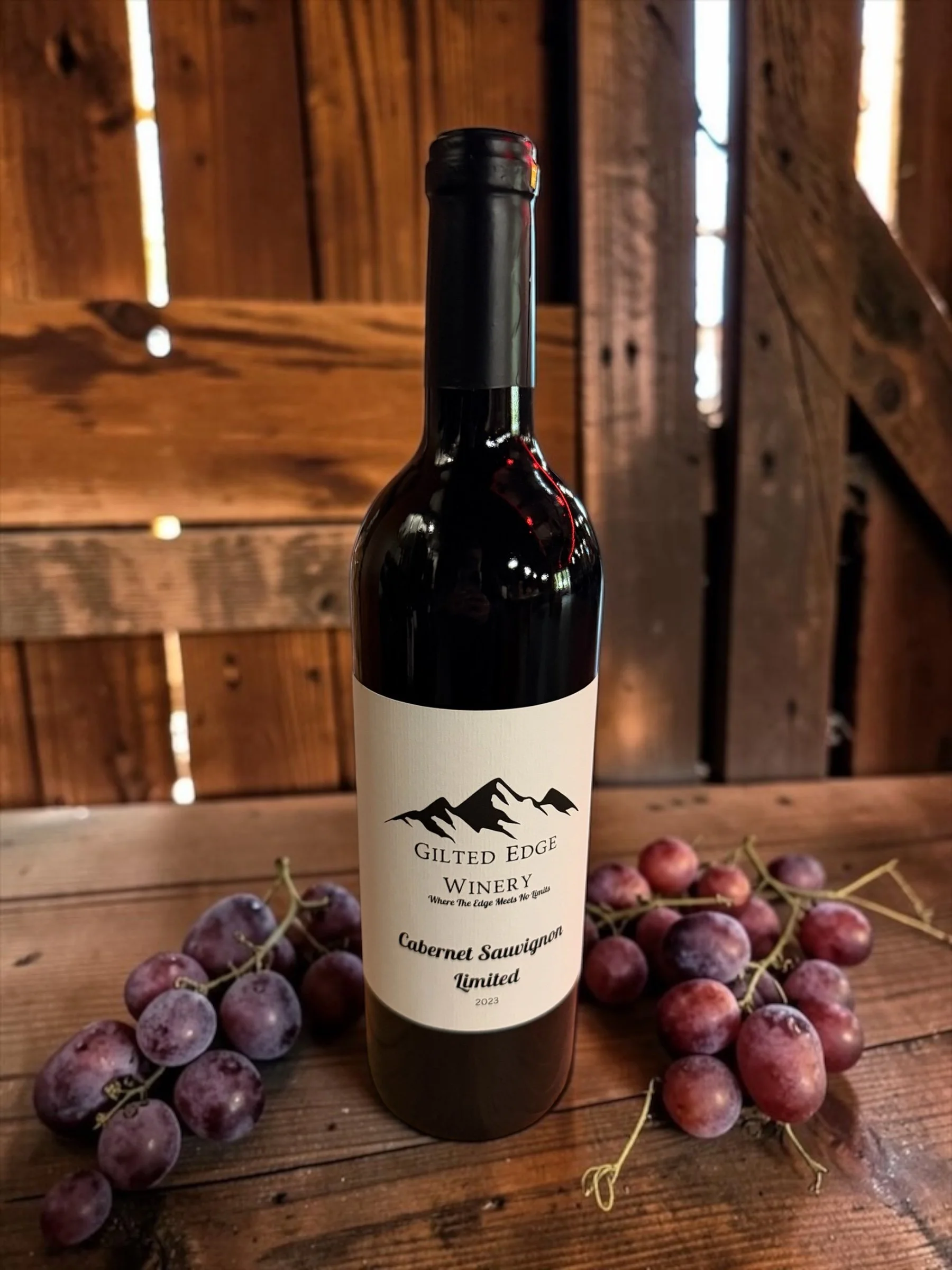 A bottle of red wine from Gilt Edge Winery surrounded by red grapes on a wooden surface, with a wooden wall in the background.