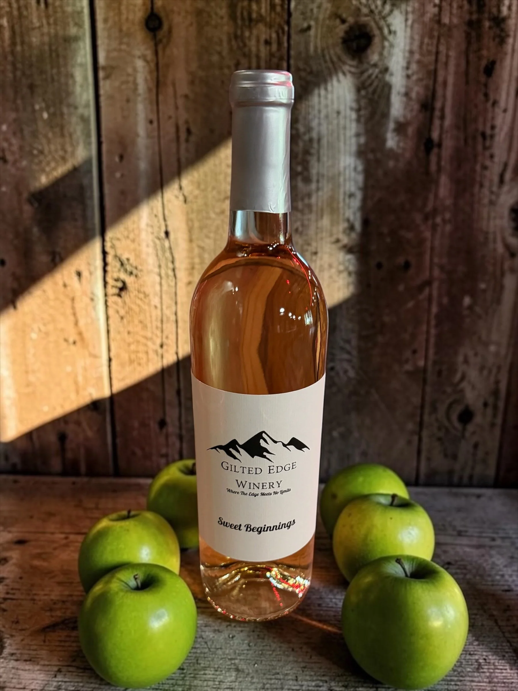 A bottle of wine from Gilded Edge Winery surrounded by green apples on a wooden surface with a wooden background.