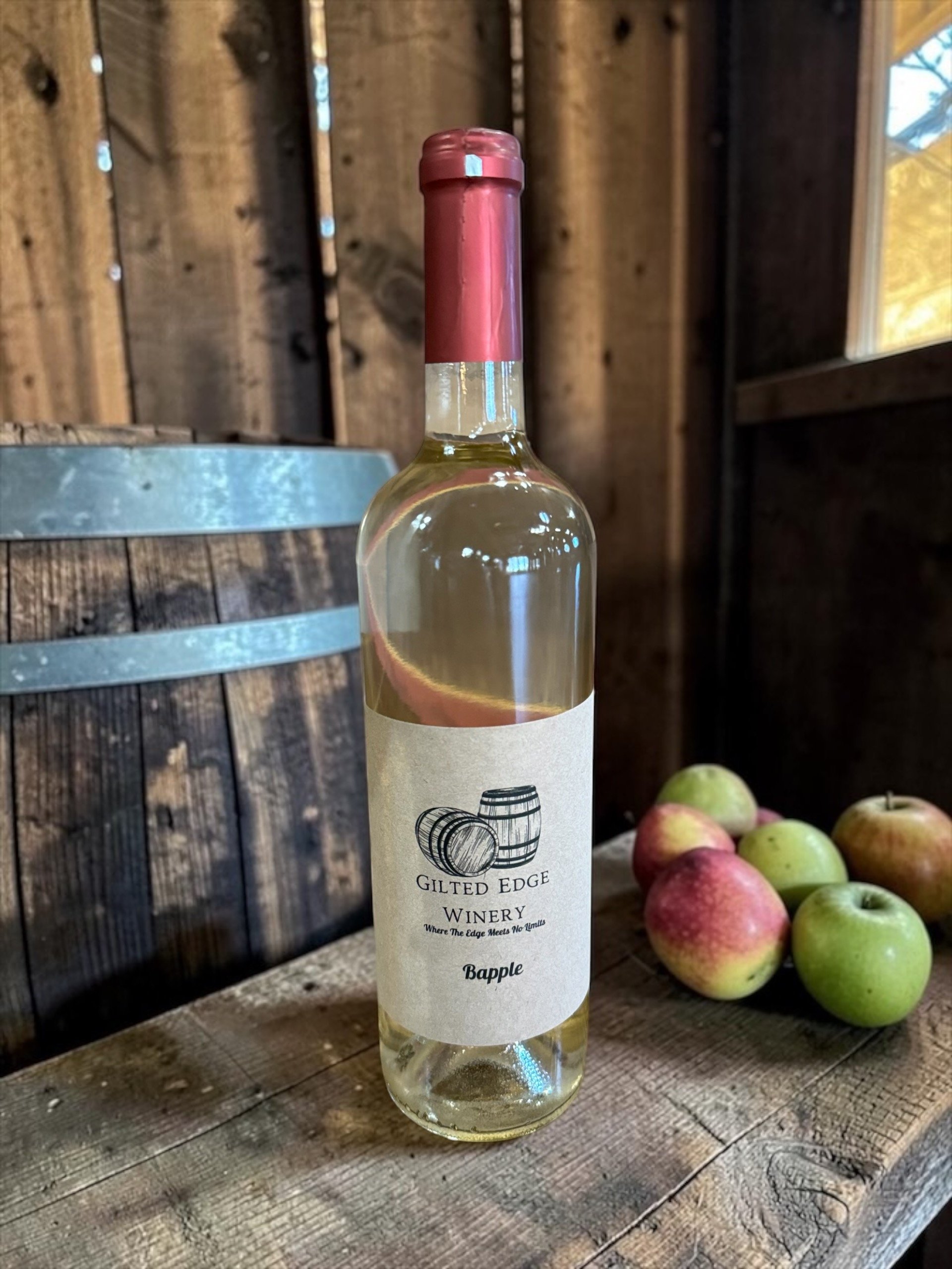 A bottle of white wine labeled 'Gilted Edge Winery' placed on a rustic wooden table next to a bunch of apples. In the background, there is a wooden barrel and wooden wall paneling with natural lighting.