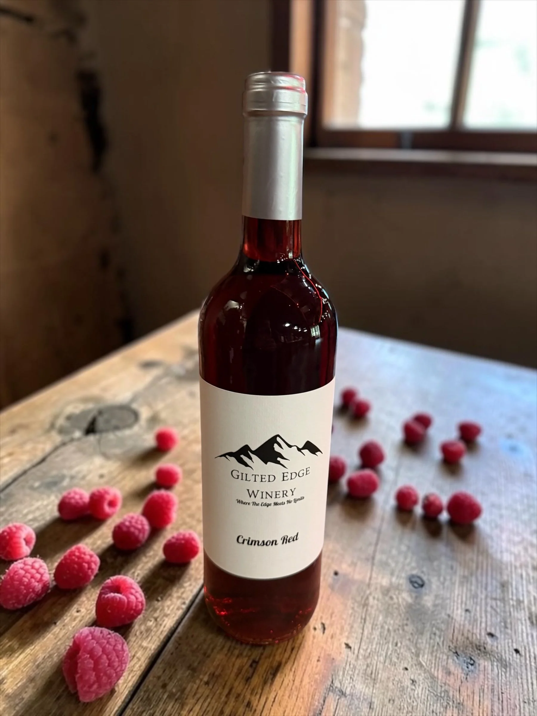 A bottle of Crimson Red wine from Gilted Edge Winery on a wooden table with scattered pink raspberries and a window in the background.