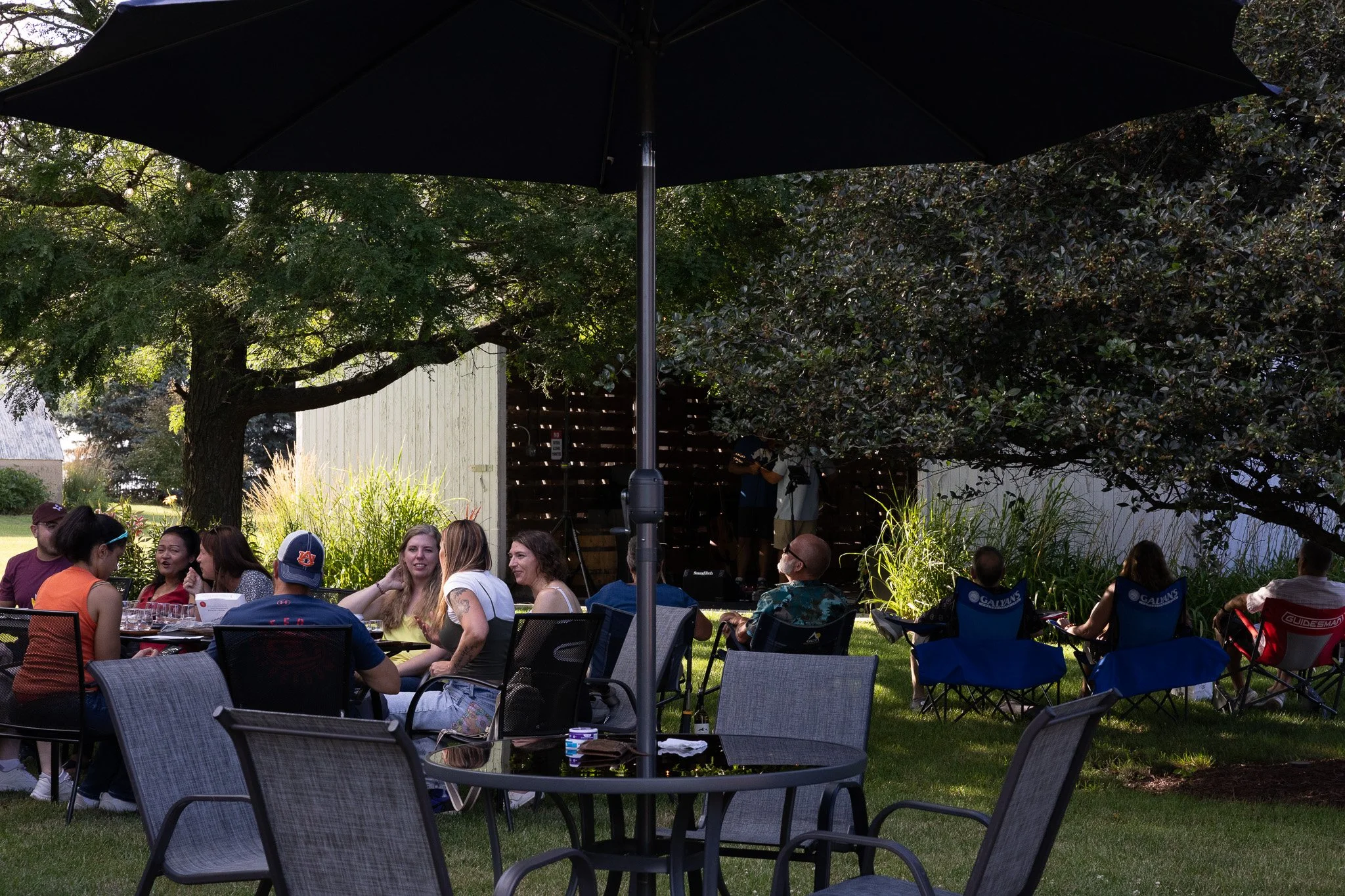People gathered outdoors under a large black umbrella, sitting at tables, with a stage and trees in the background during a daytime event.