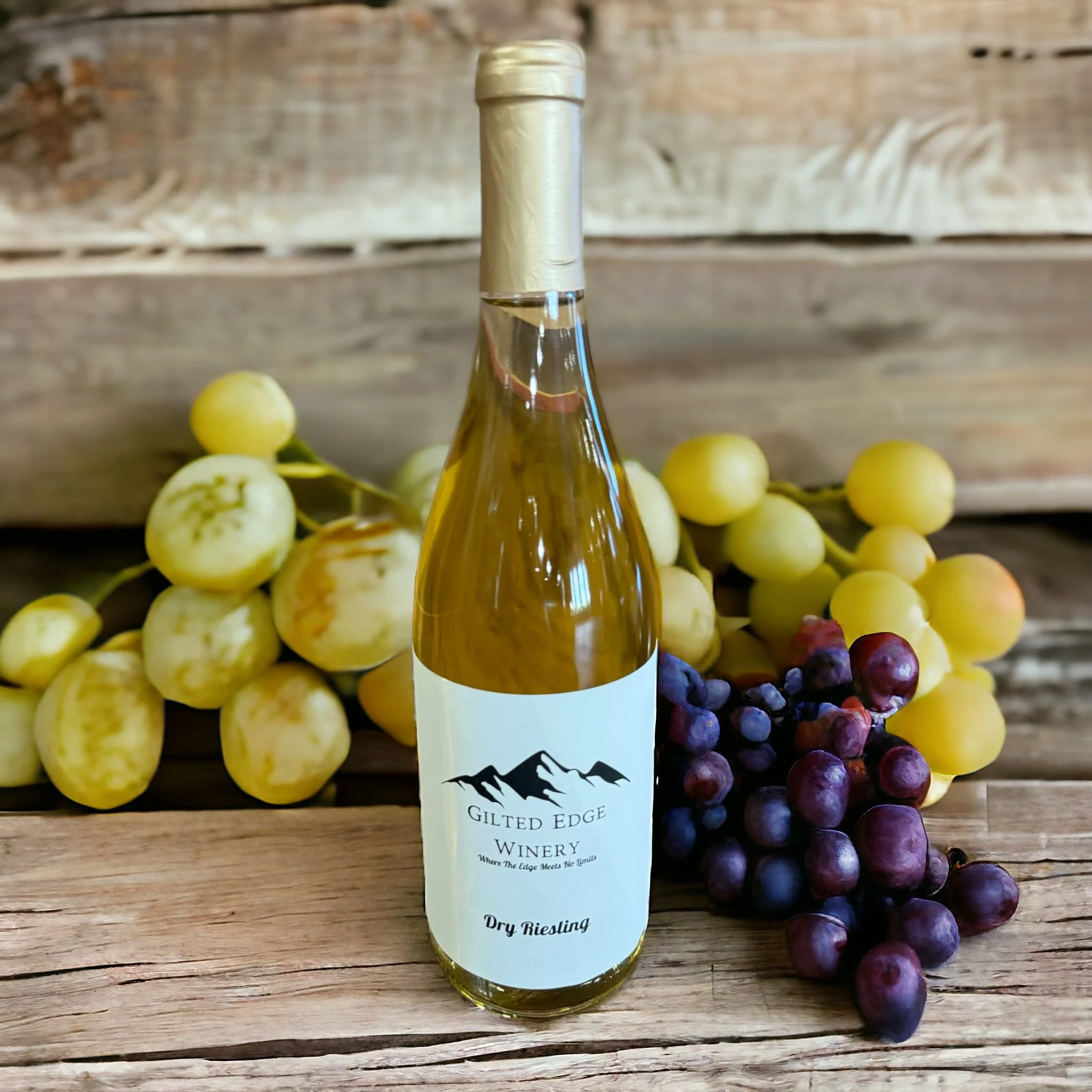 A bottle of dry Riesling wine from Gilt Edge Winery surrounded by green and purple grapes on a wooden surface.