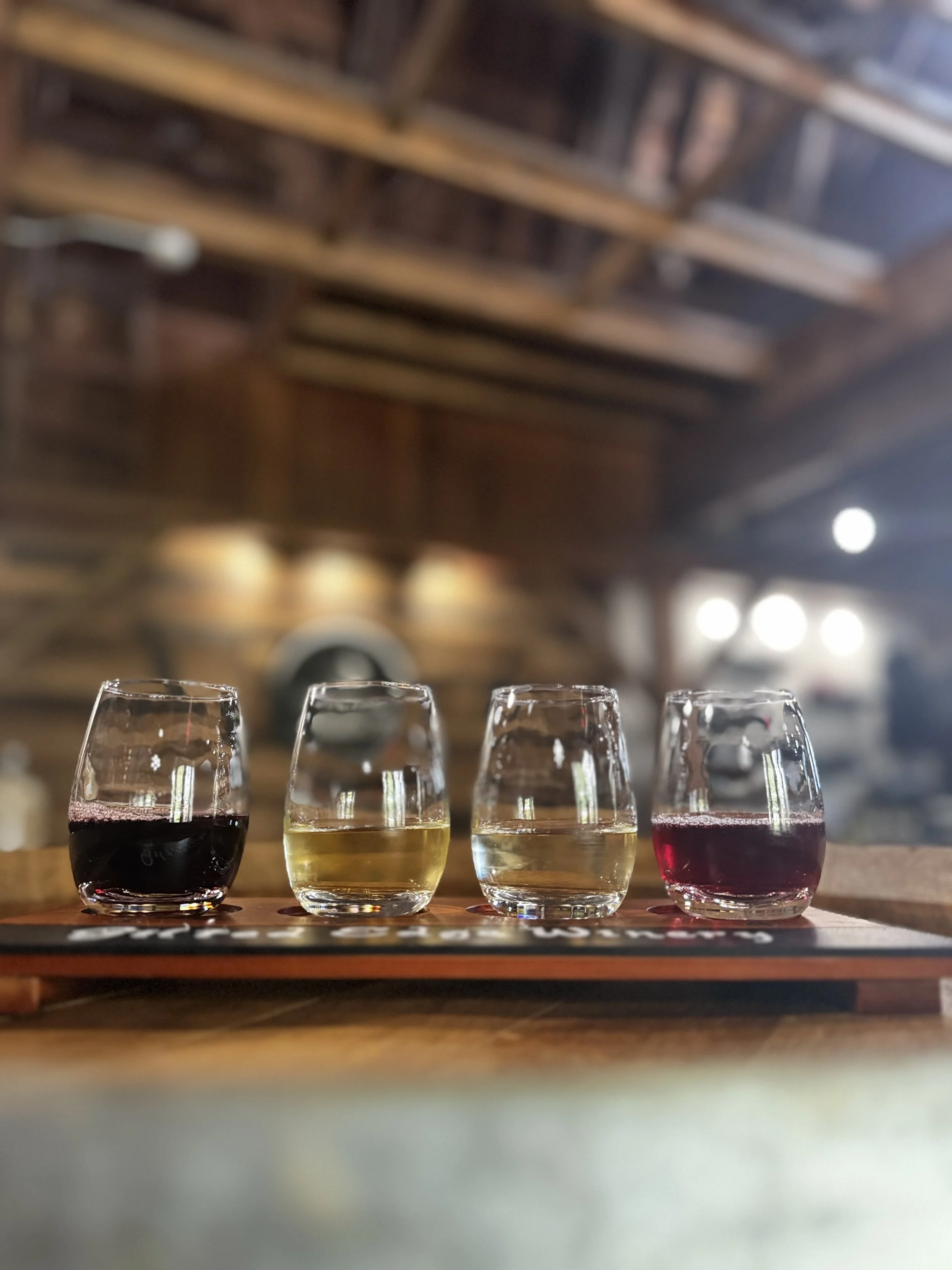Four glasses of wine in different colors—red, white, and rosé—placed on a wooden table in a bar or winery setting with wine barrels and ceiling beams in the background.