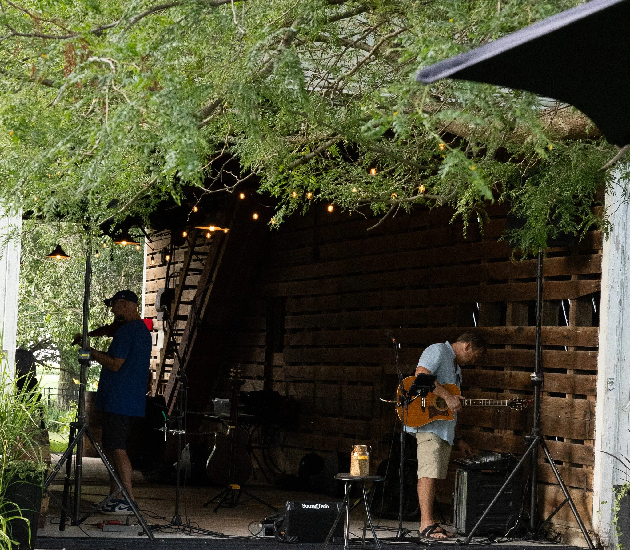 Two men performing music on an outdoor stage under trees. One playing a guitar, the other adjusting equipment, with string lights hanging in the background.