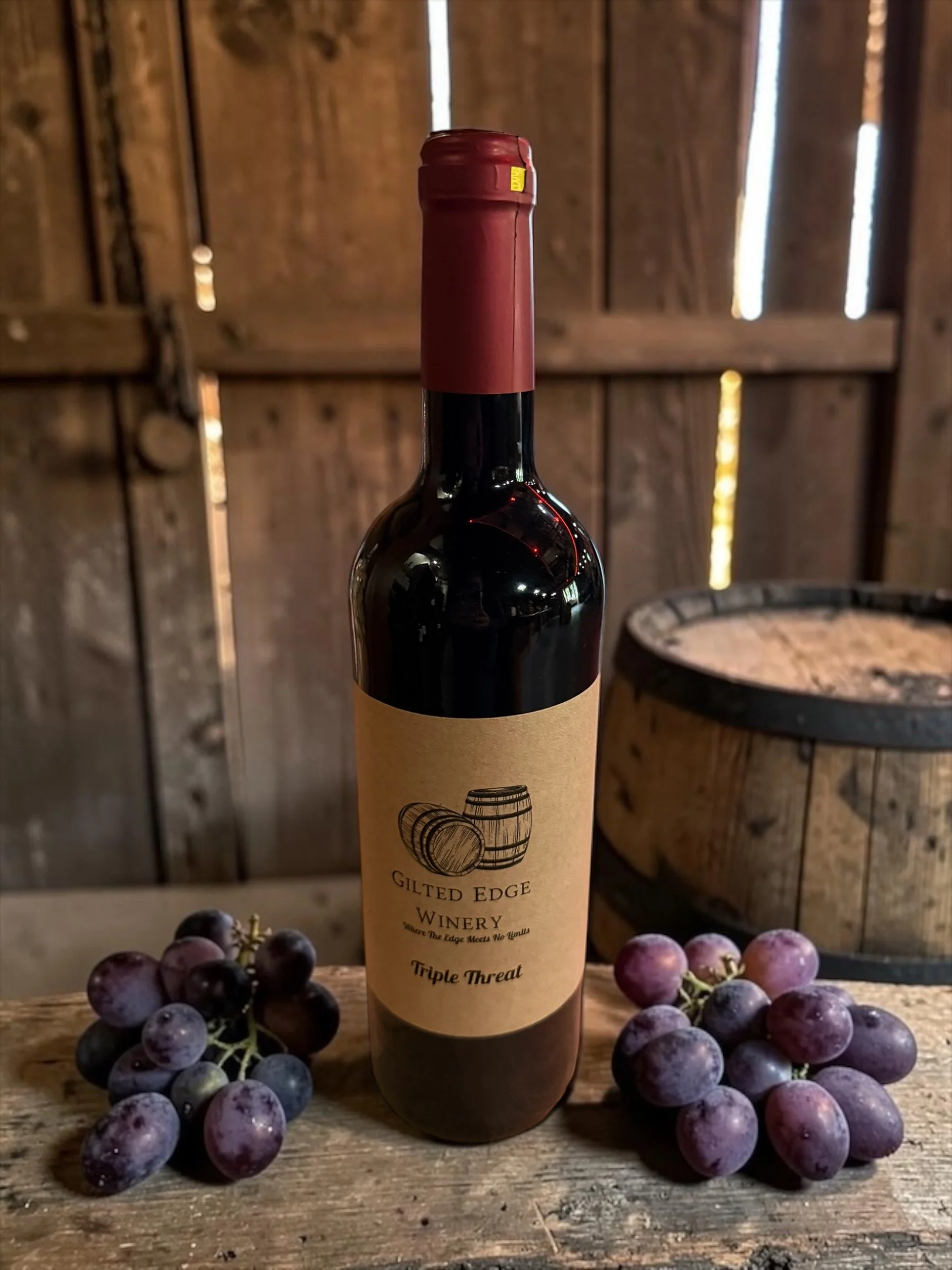 A bottle of red wine labeled 'Triple Threat' from Gilded Edge Winery, surrounded by bunches of purple grapes on a wooden surface, with a rustic wooden background.